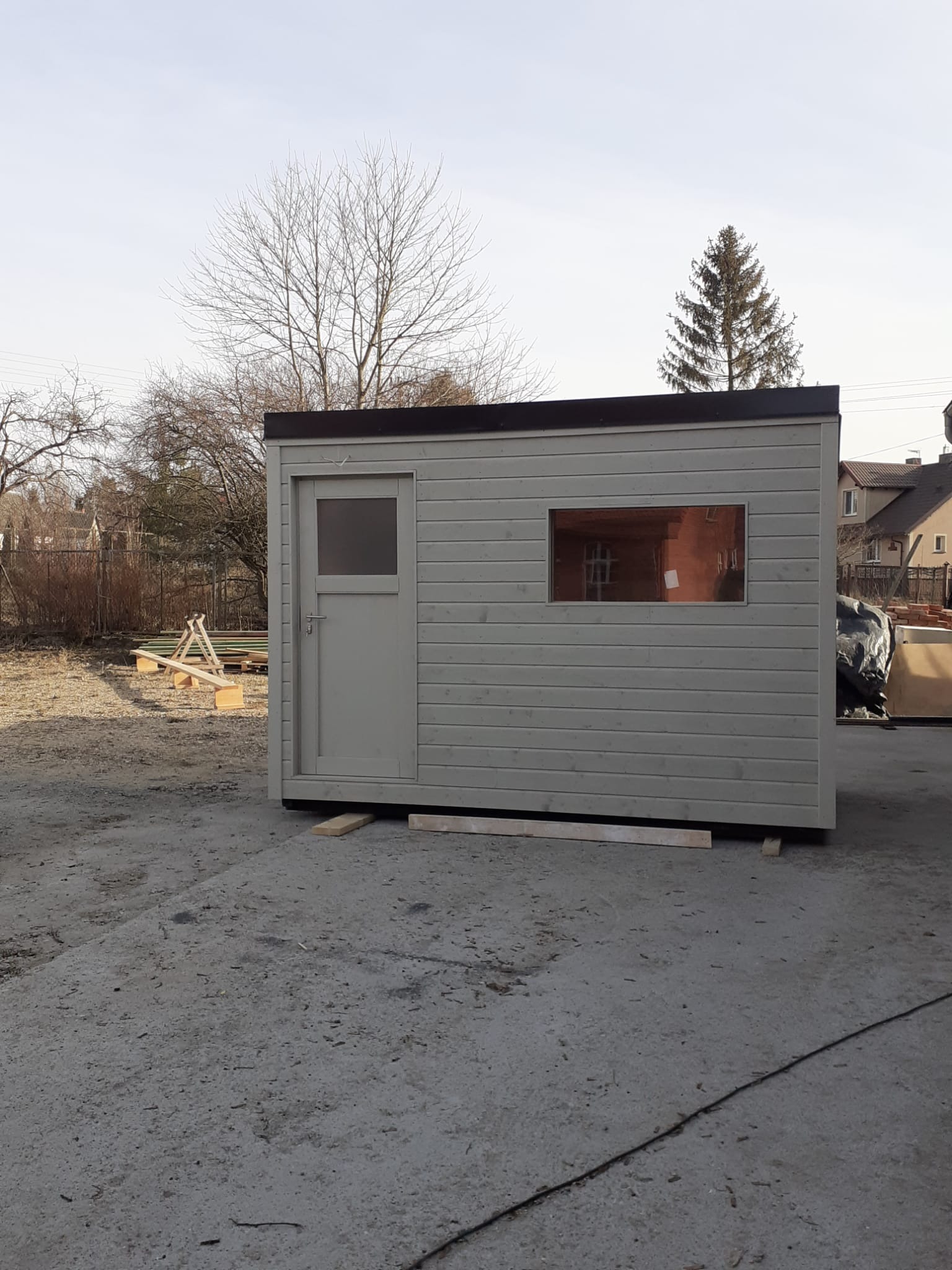 Nowoczesna, szara sauna ogrodowa z oknem i drzwiami, ustawiona na betonowej powierzchni. W tle drzewa i budynki mieszkalne.