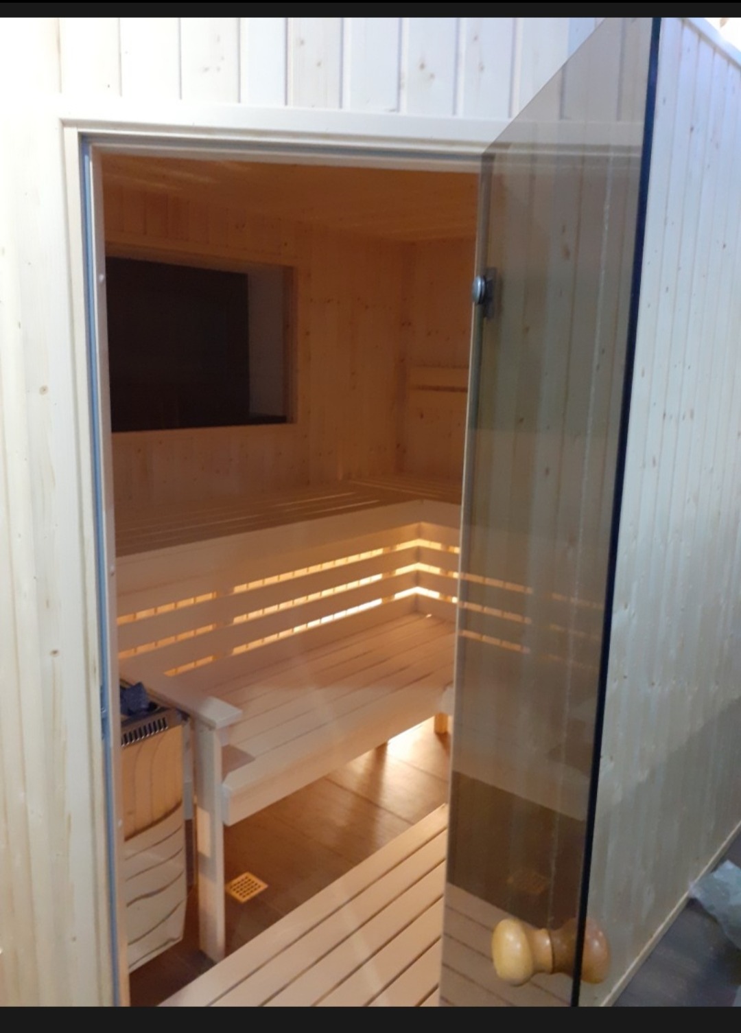 Jasne drewno w saunie z przeszklonymi drzwiami. Widoczne ławki i piec. Ujęcie z zewnątrz, podkreślające estetykę i funkcjonalność.