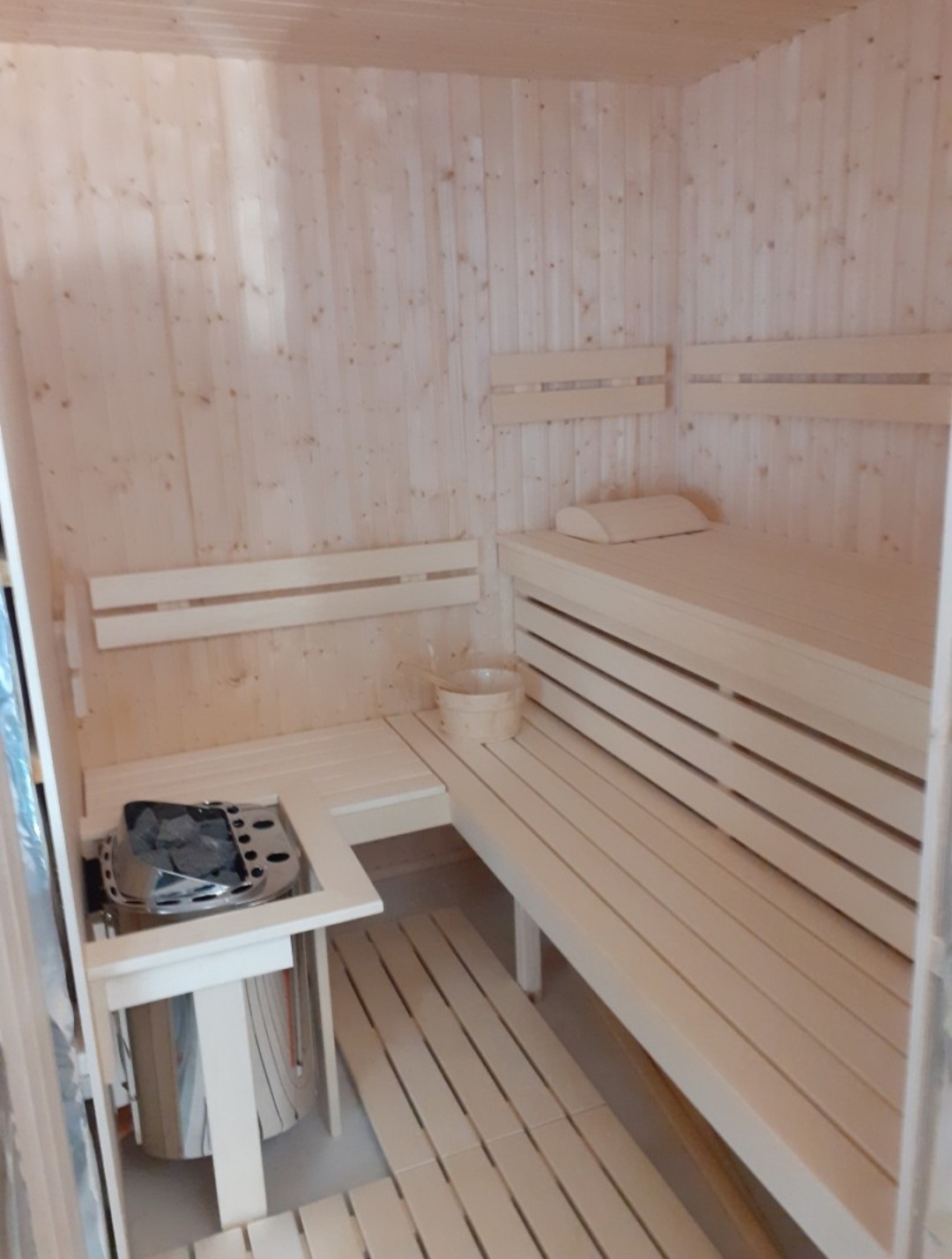 Jasne wnętrze sauny z drewnianymi ławkami, piecem i wiadrem. Widok na piec z kamieniami i drewniane wykończenie ścian. Minimalistyczny design.