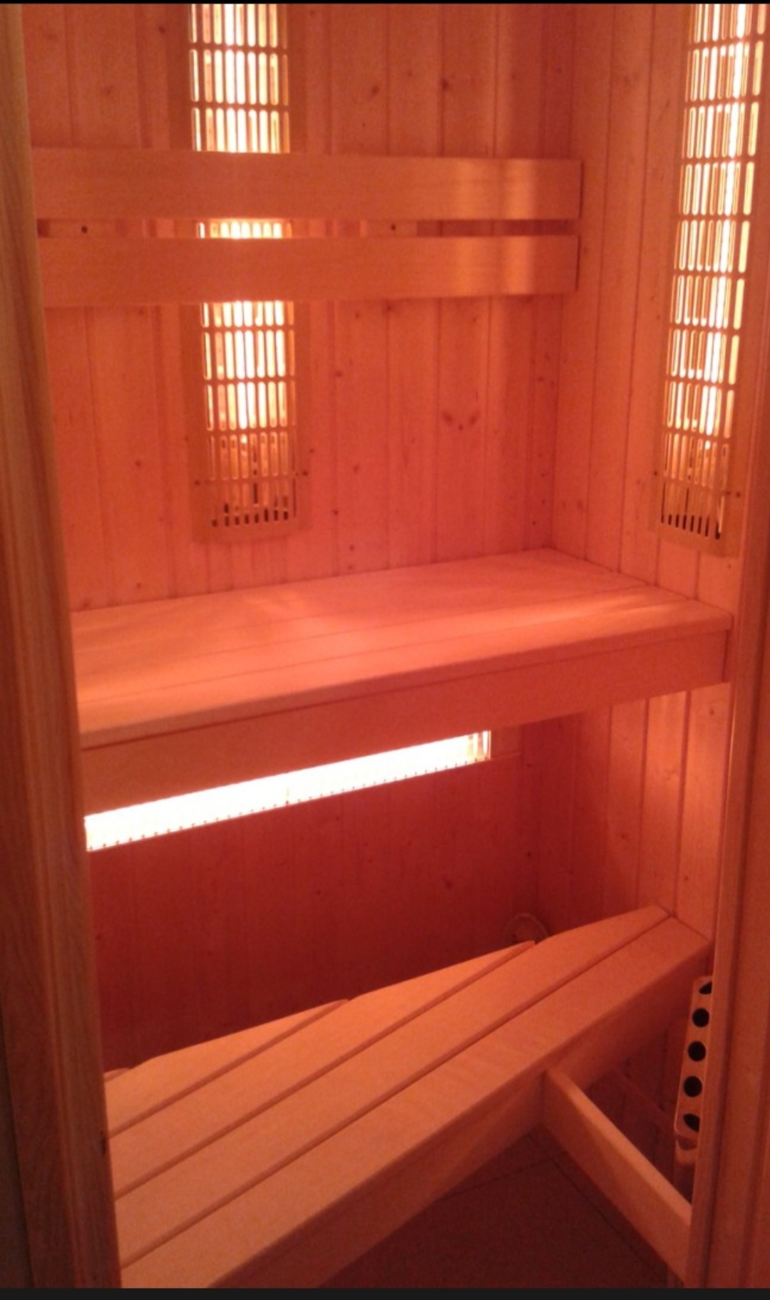 Wnętrze sauny z jasnego drewna, dwupoziomowe ławki, oświetlenie punktowe, ciepła kolorystyka, widok z perspektywy wejścia, minimalistyczny design.