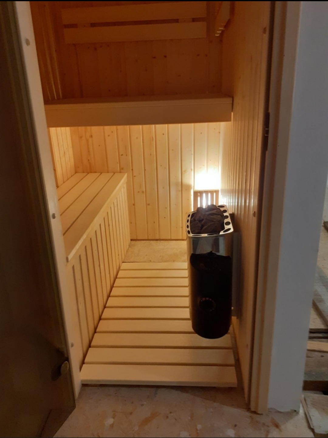 Wnętrze sauny z drewnianymi ławkami i piecem, widok z zewnątrz przez otwarte drzwi. Jasne drewno, ciepłe oświetlenie, minimalistyczny design.