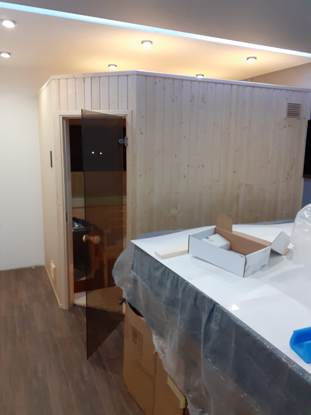 Jasna, drewniana sauna z przeszklonymi drzwiami, widoczna z boku. Wnętrze sauny oświetlone, na pierwszym planie biały blat z akcesoriami.