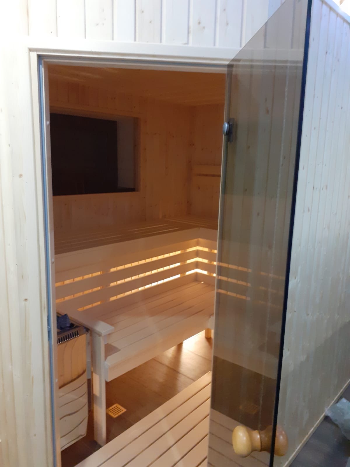 Jasne drewno i szklane drzwi w saunie z ławkami, widok z zewnątrz. Minimalistyczny design, ciepłe oświetlenie, widoczny piec i kratka wentylacyjna.