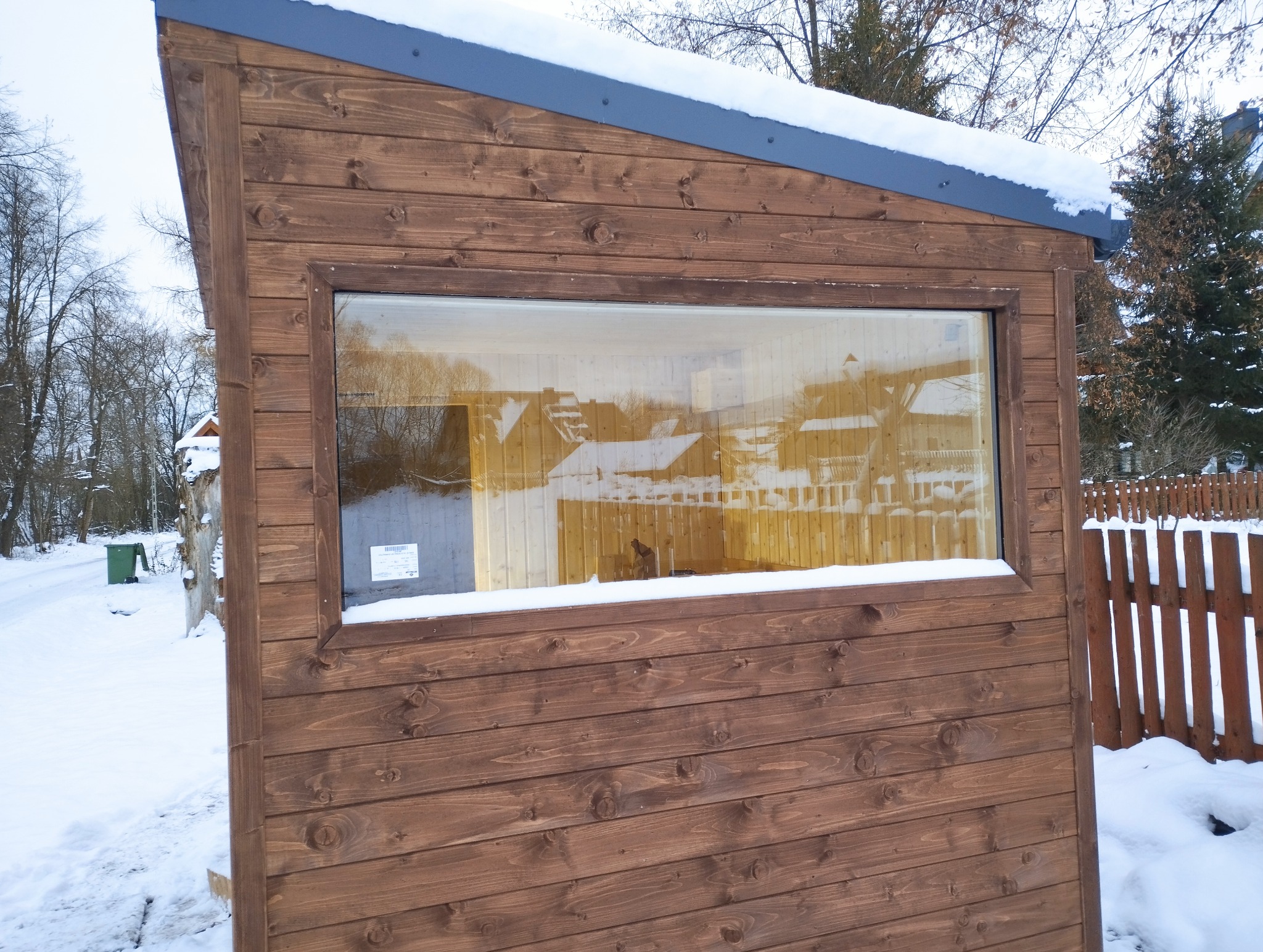 Drewniana konstrukcja z oknem w zimowym krajobrazie. Widoczne deski z wyraźnym usłojeniem i śnieg na dachu. W oknie odbicie zabudowań.