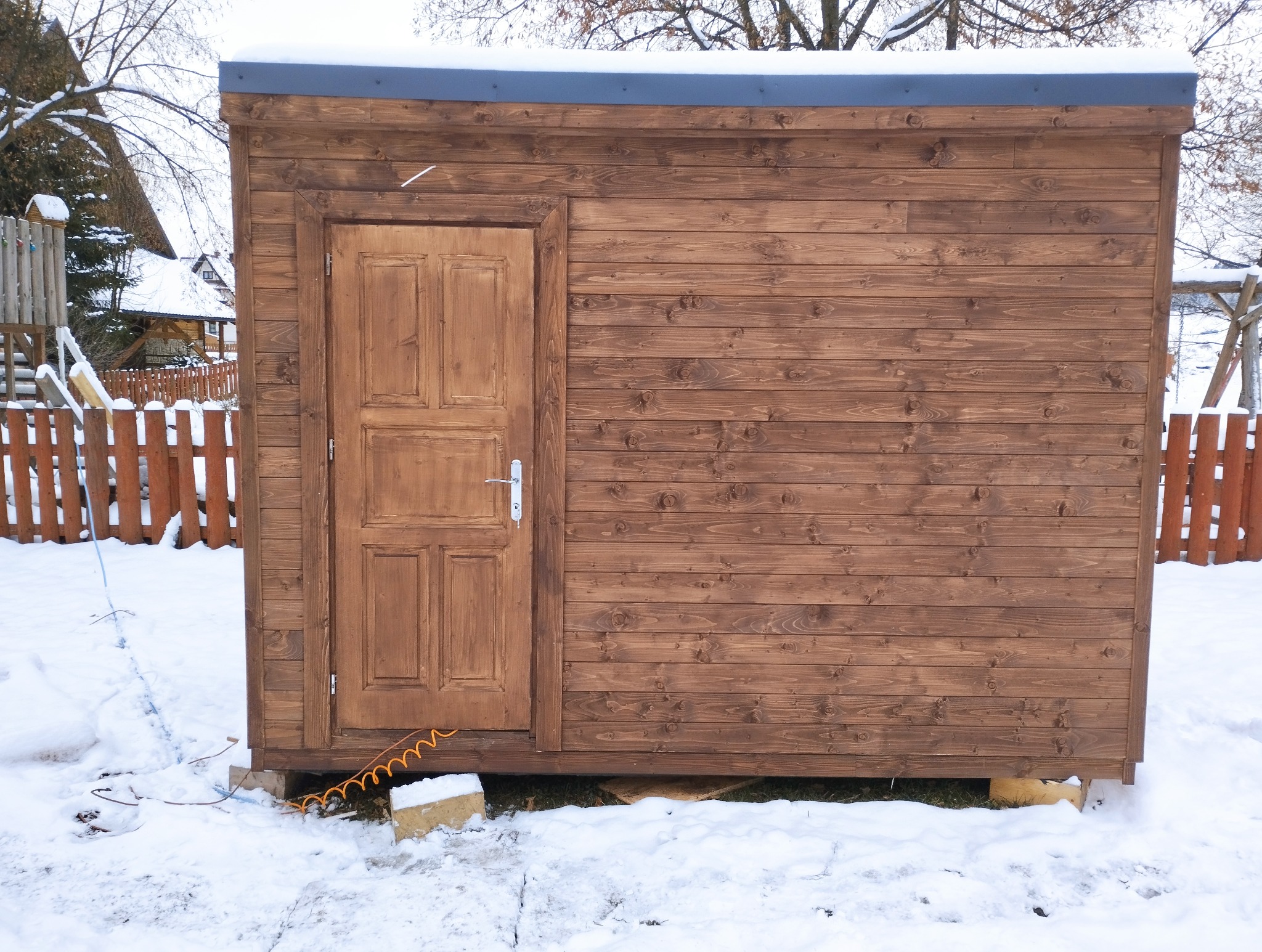 Drewniana, brązowa sauna ogrodowa z drzwiami, widoczna zimą na tle zasypanego śniegiem ogrodu. Konstrukcja z desek, z szarym dachem i widocznymi elementami instalacji.