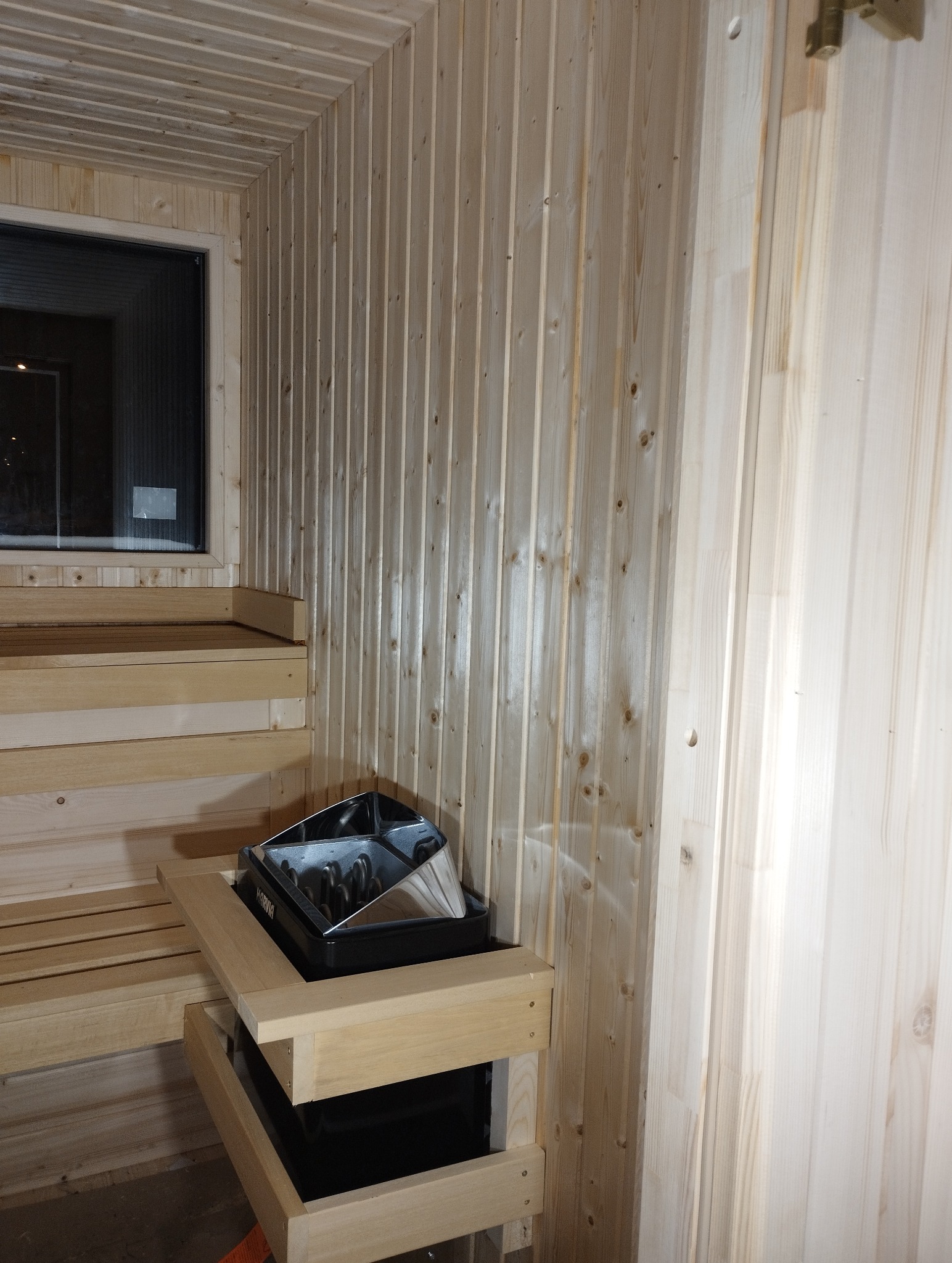 Jasne drewno w saunie: ławy, ściany z boazerii i piec Harvia. Minimalistyczny design, ciepłe oświetlenie, widok na zewnątrz przez okno.