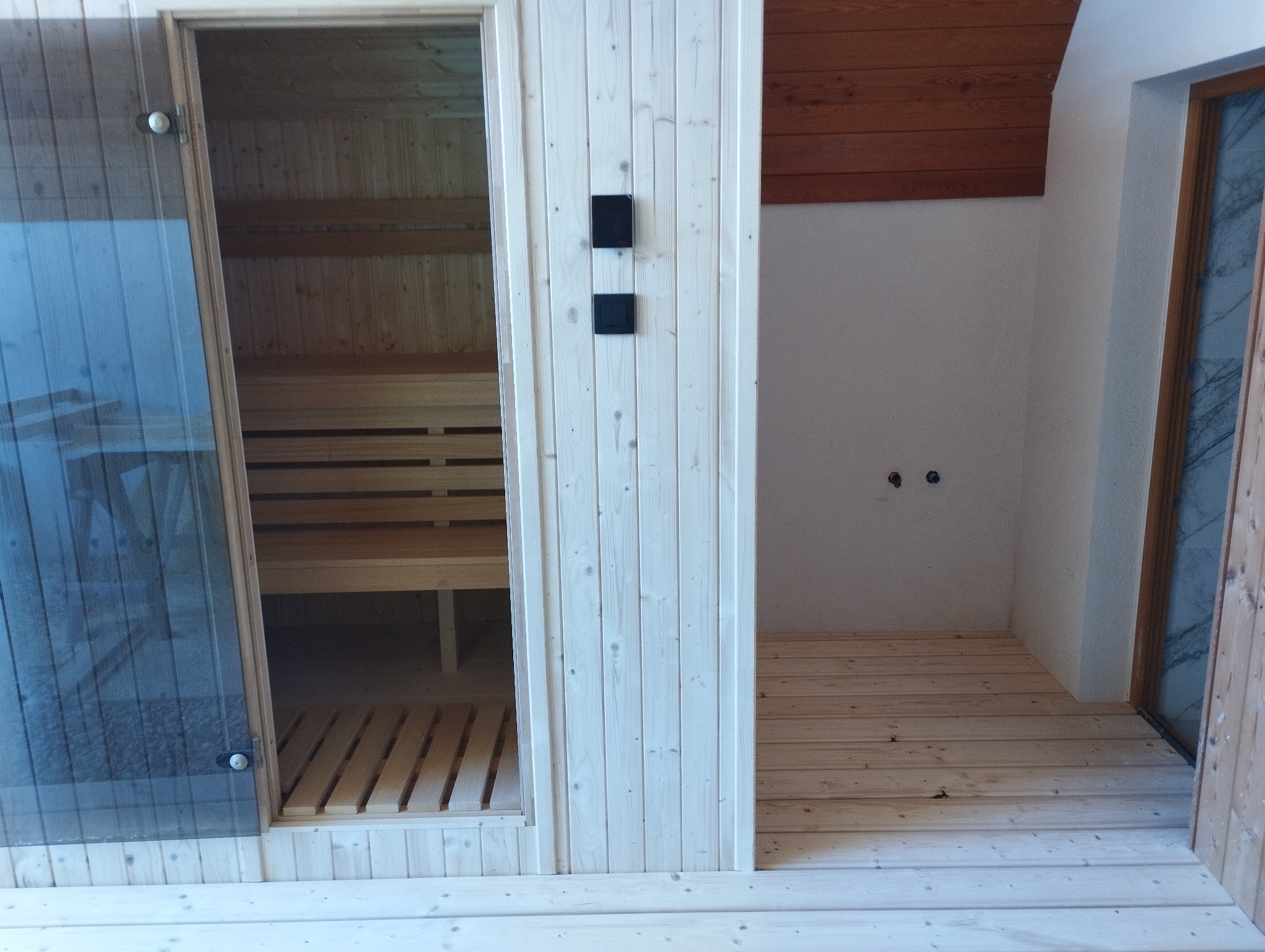 Wnętrze sauny z drewnianymi ławkami, widoczne przez szklane drzwi. Obok pomieszczenie z drewnianą podłogą i białymi ścianami, z widocznymi przyłączami.