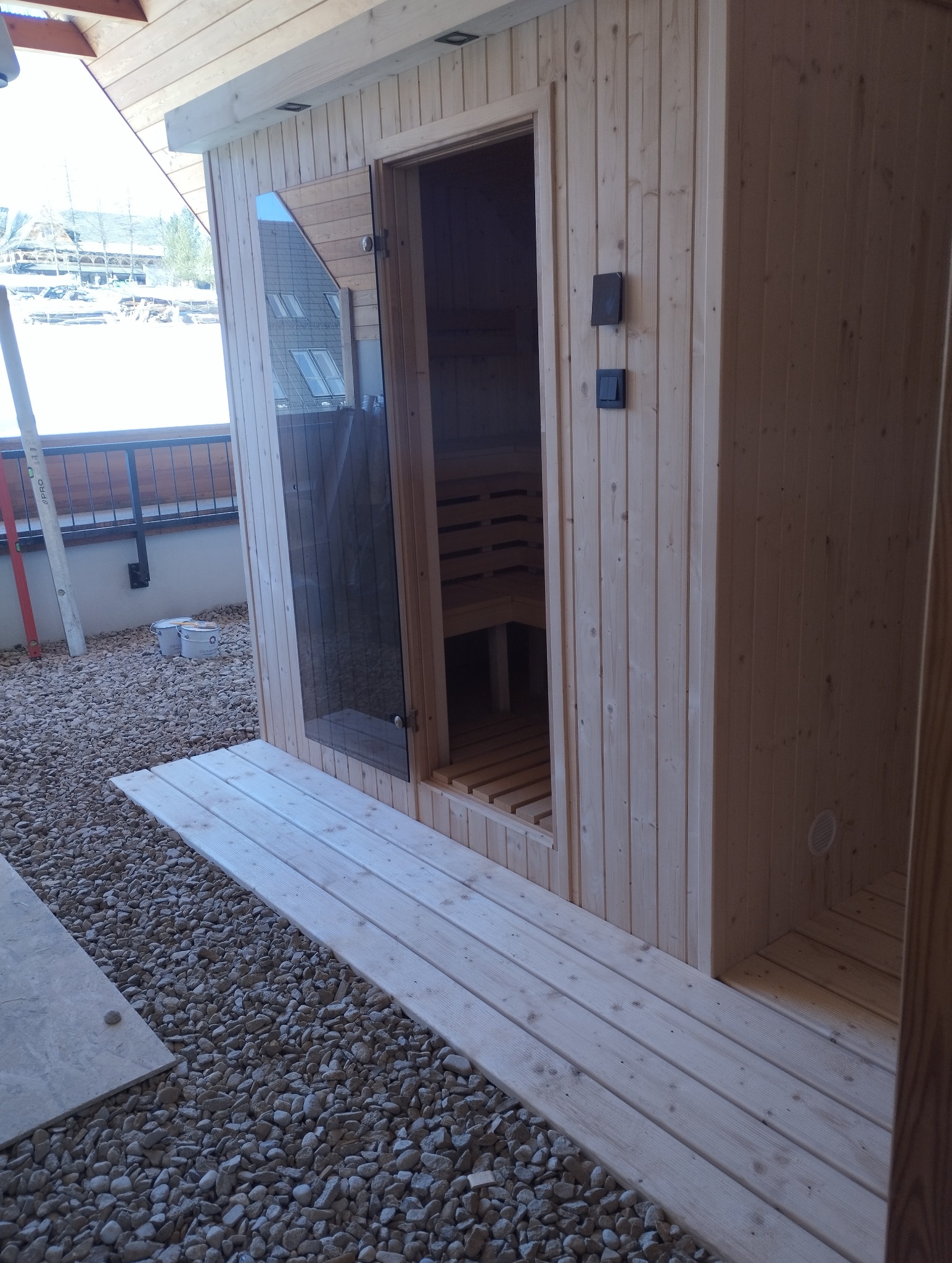 Drewniana sauna zewnętrzna z przeszklonymi drzwiami, umieszczona na żwirowym podłożu z drewnianym podejściem. Widoczne wnętrze sauny z ławkami.
