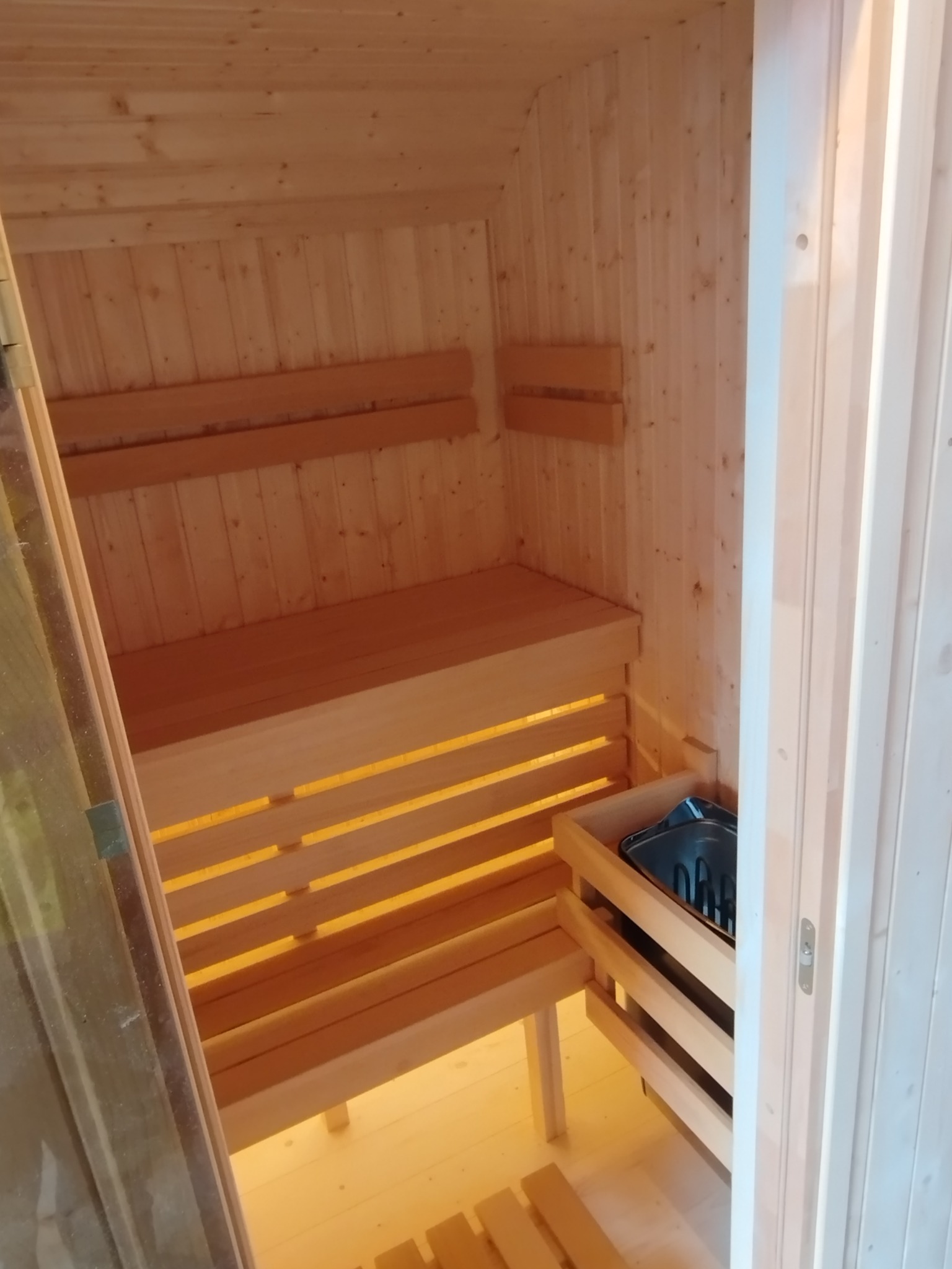 Jasne drewno w saunie: ławy, ściany i piec z kamieniami. Widok zza szklanych drzwi, ciepłe oświetlenie podkreśla naturalny rysunek drewna.