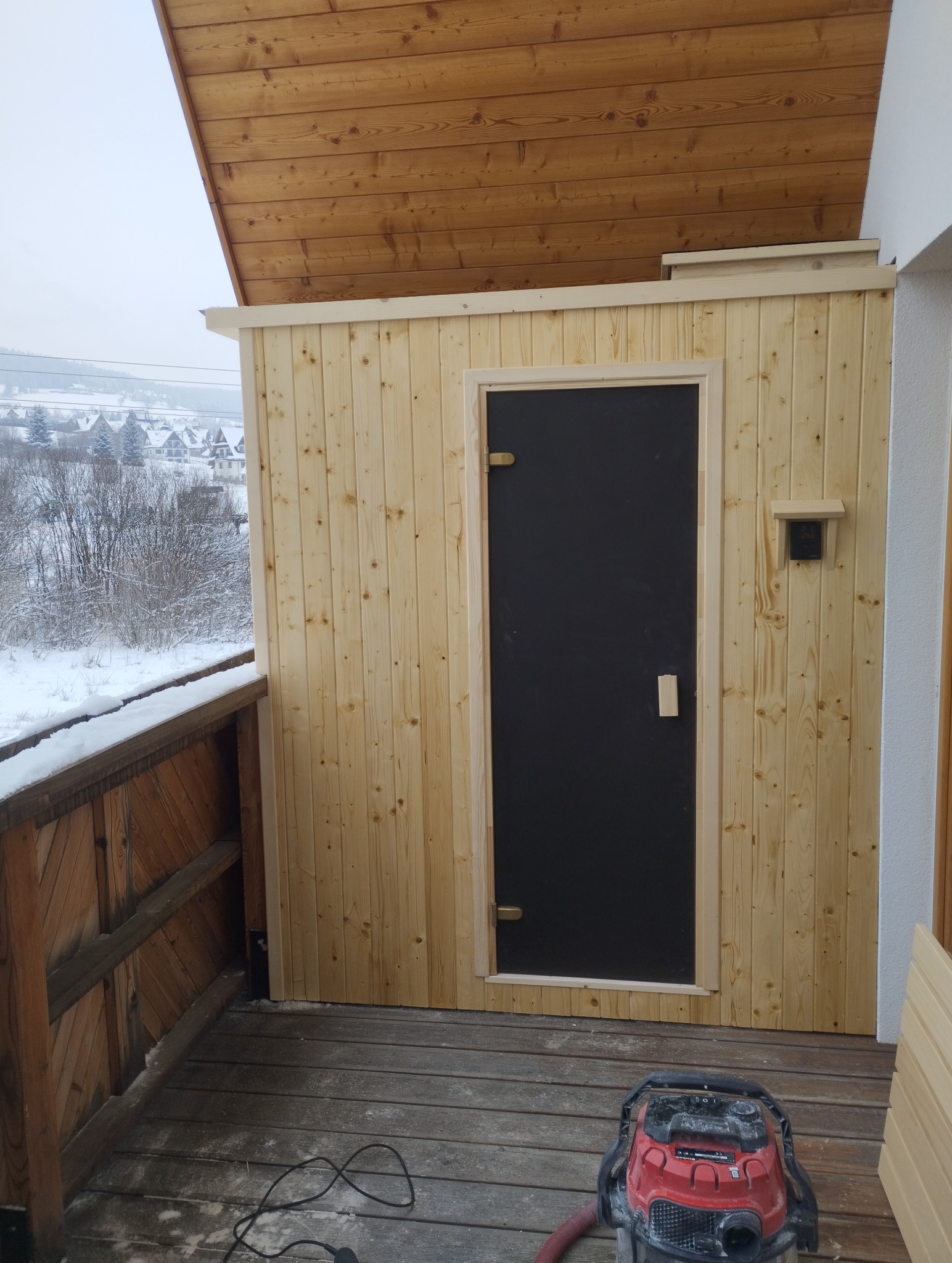 Drewniana sauna na balkonie z czarnymi drzwiami i widokiem na zimowy krajobraz. Na podłodze widoczny odkurzacz przemysłowy.