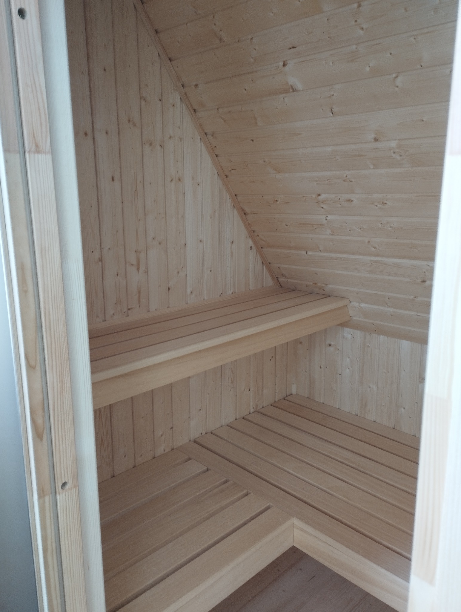 Wnętrze sauny z jasnego drewna, widok z perspektywy wejścia. Ukośny sufit, drewniane ławki i podłoga z desek tworzą minimalistyczny, skandynawski design.