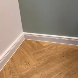 Fragment wnętrza: podłoga wyłożona panelami imitującymi drewno ułożonymi we wzór jodełki, białe listwy przypodłogowe, ściany pomalowane na biało i szaro-zielono.