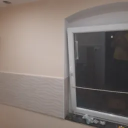 Ściana pomalowana na beżowo, z dekoracyjnymi płytkami ceramicznymi u dołu, okno z plastikową ramą, na parapecie szklanka i resztki po montażu płytek.