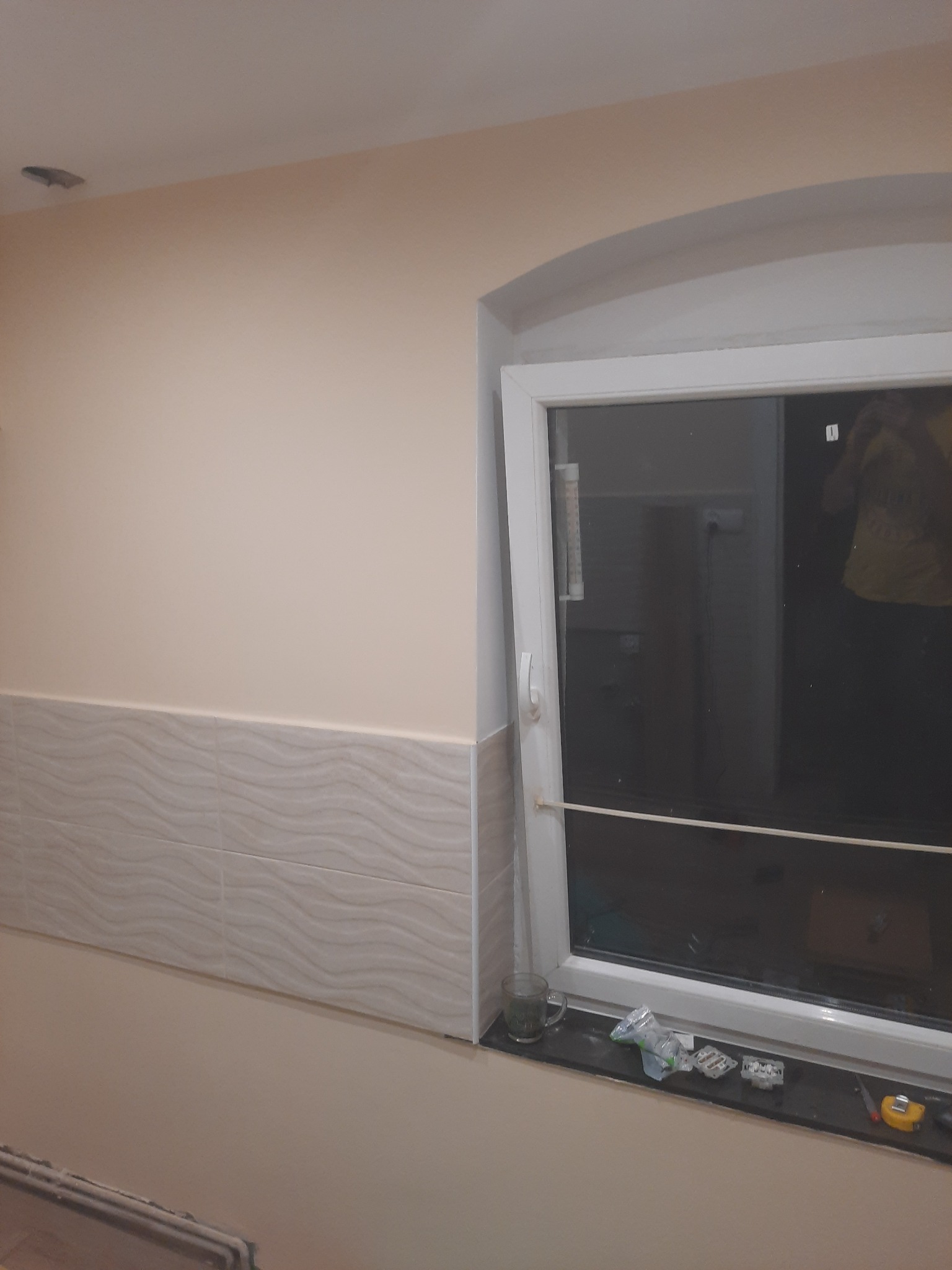 Ściana pomalowana na beżowo, z dekoracyjnymi płytkami ceramicznymi u dołu, okno z plastikową ramą, na parapecie szklanka i resztki po montażu płytek.