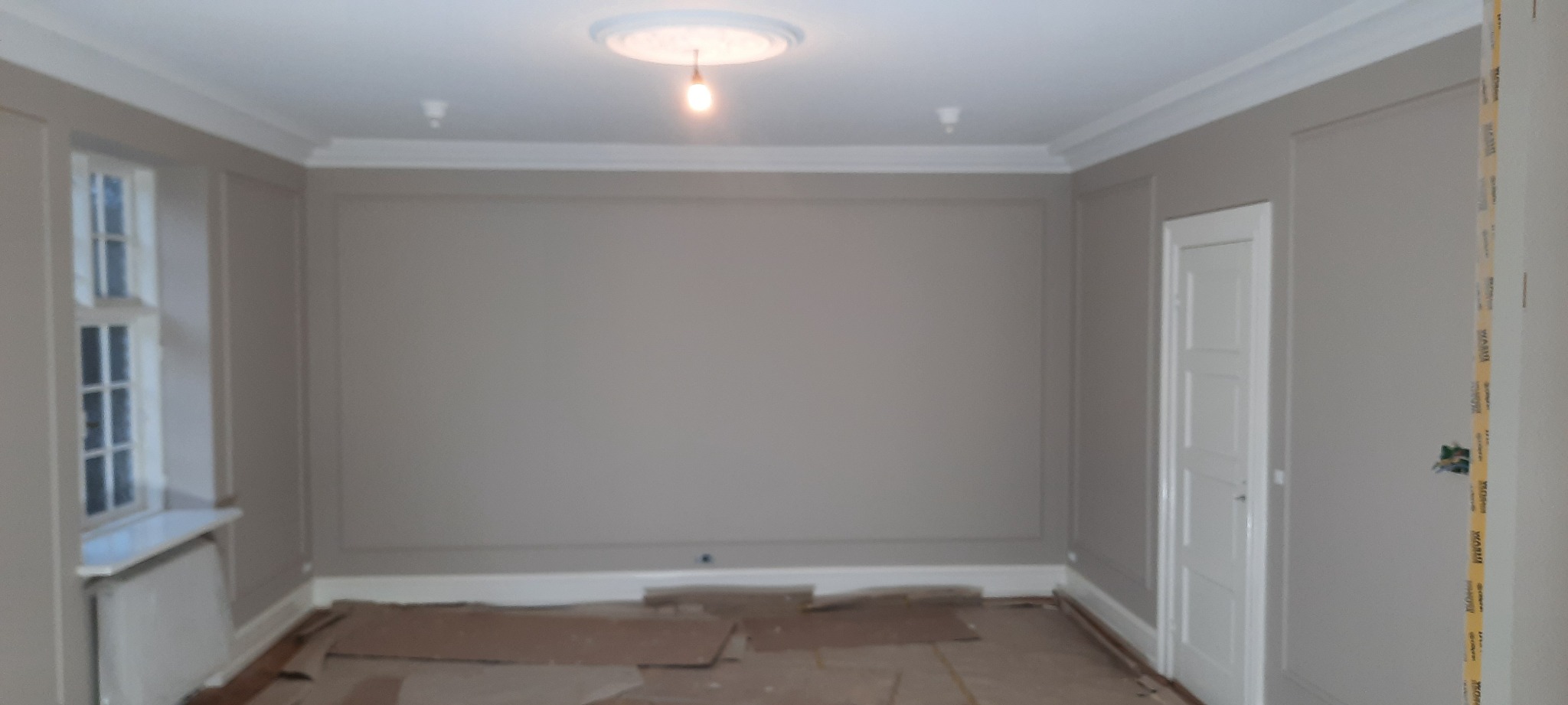 Pokój po malowaniu ścian na szaro z białymi listwami przypodłogowymi i sufitowymi, zabezpieczony kartonem na podłodze, z widoczną lampą sufitową i oknem.