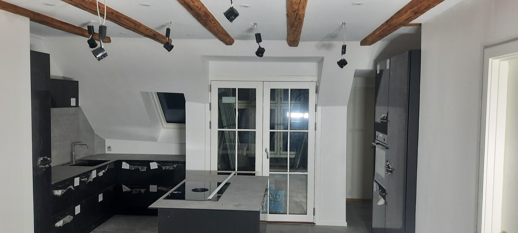 Wnętrze kuchni w trakcie remontu: surowe drewniane belki z oświetleniem punktowym, czarne szafki kuchenne częściowo zabezpieczone folią, szare blaty, okno dachowe, białe dwuskrzydłowe drzwi...