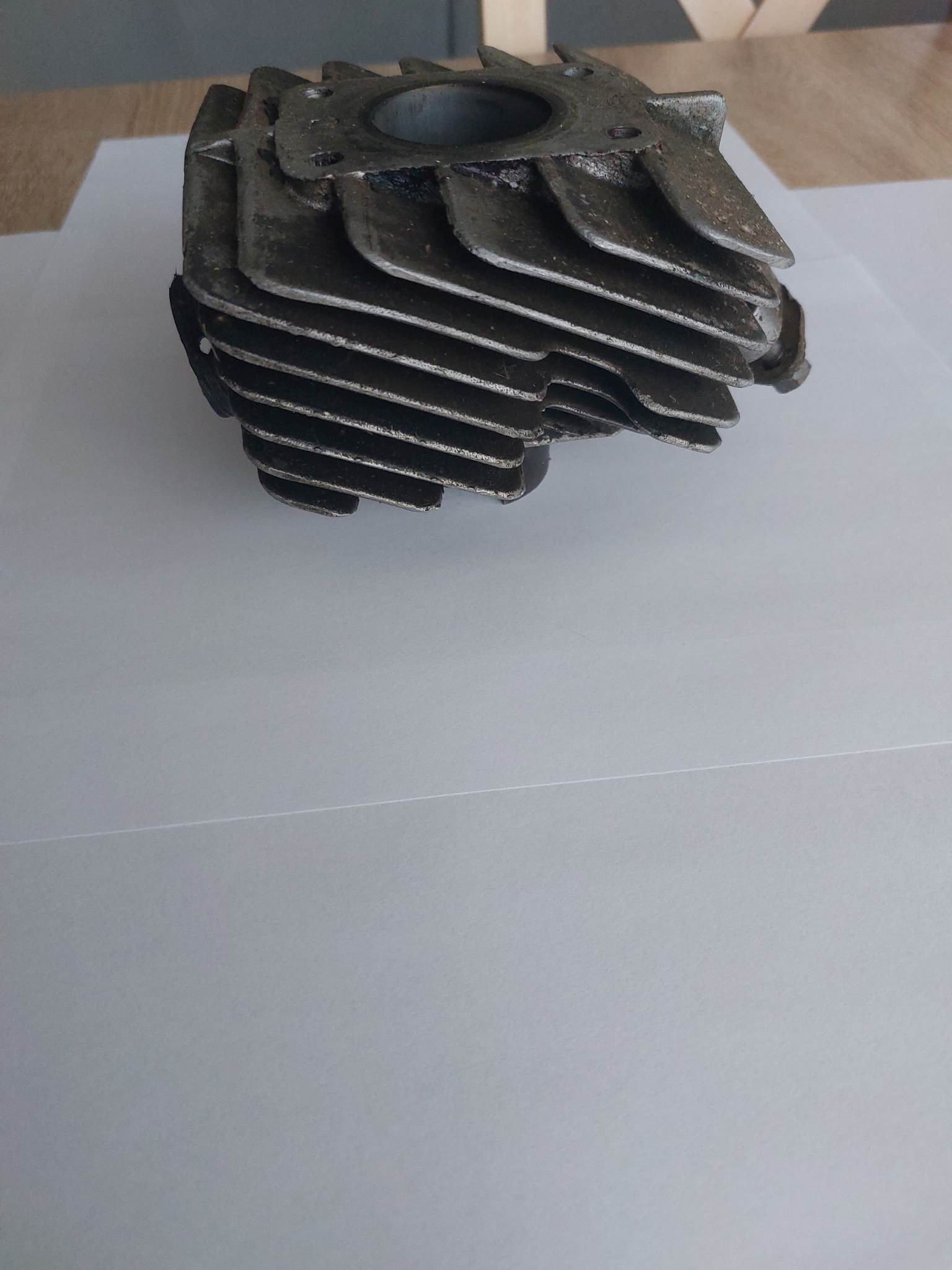 Żeliwny cylinder silnika spalinowego z żebrami chłodzącymi, widoczne ślady korozji i zabrudzeń, leżący na białym papierze.