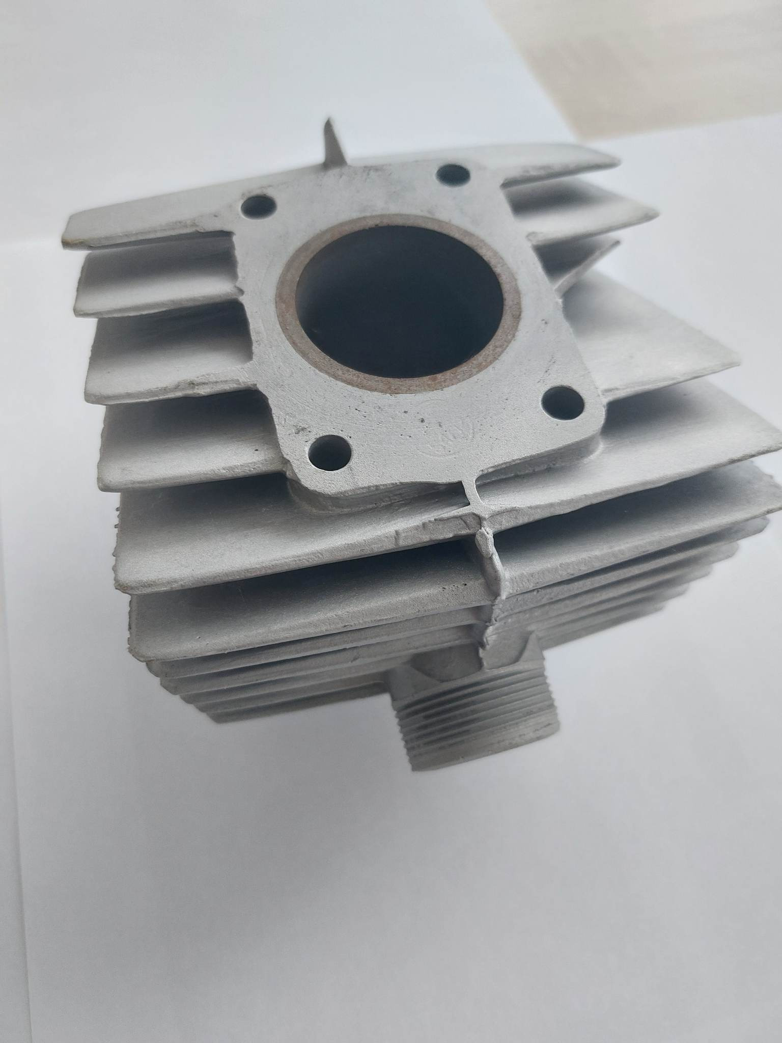 Szary, surowy odlew bloku silnika z widocznymi żebrami chłodzącymi i gwintowanym otworem, sfotografowany na białym tle.