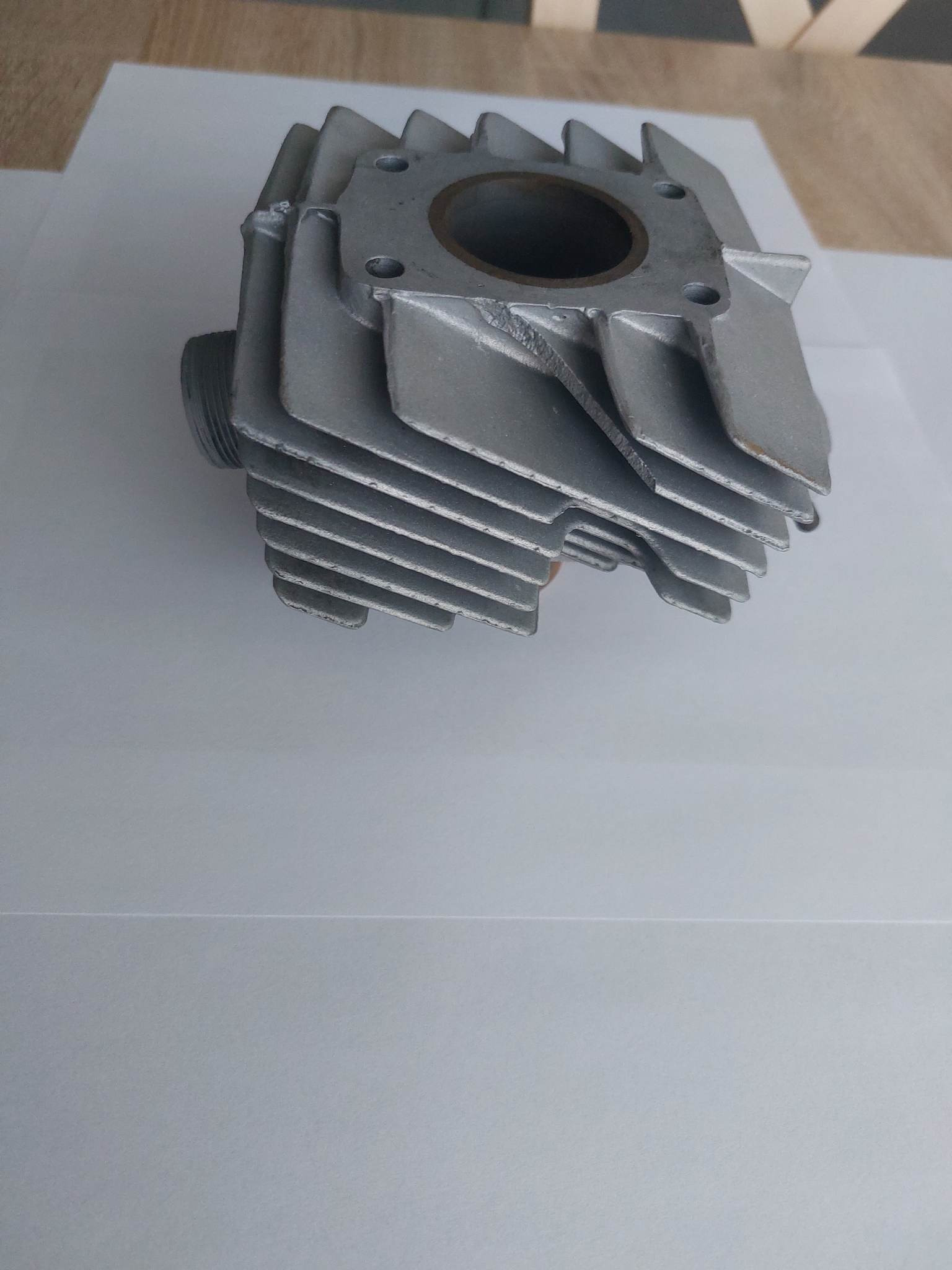 Szary cylinder silnika motocyklowego z wyraźnymi żeberkami chłodzącymi, leżący na białym tle z widocznym drobnym uszkodzeniem na krawędzi.