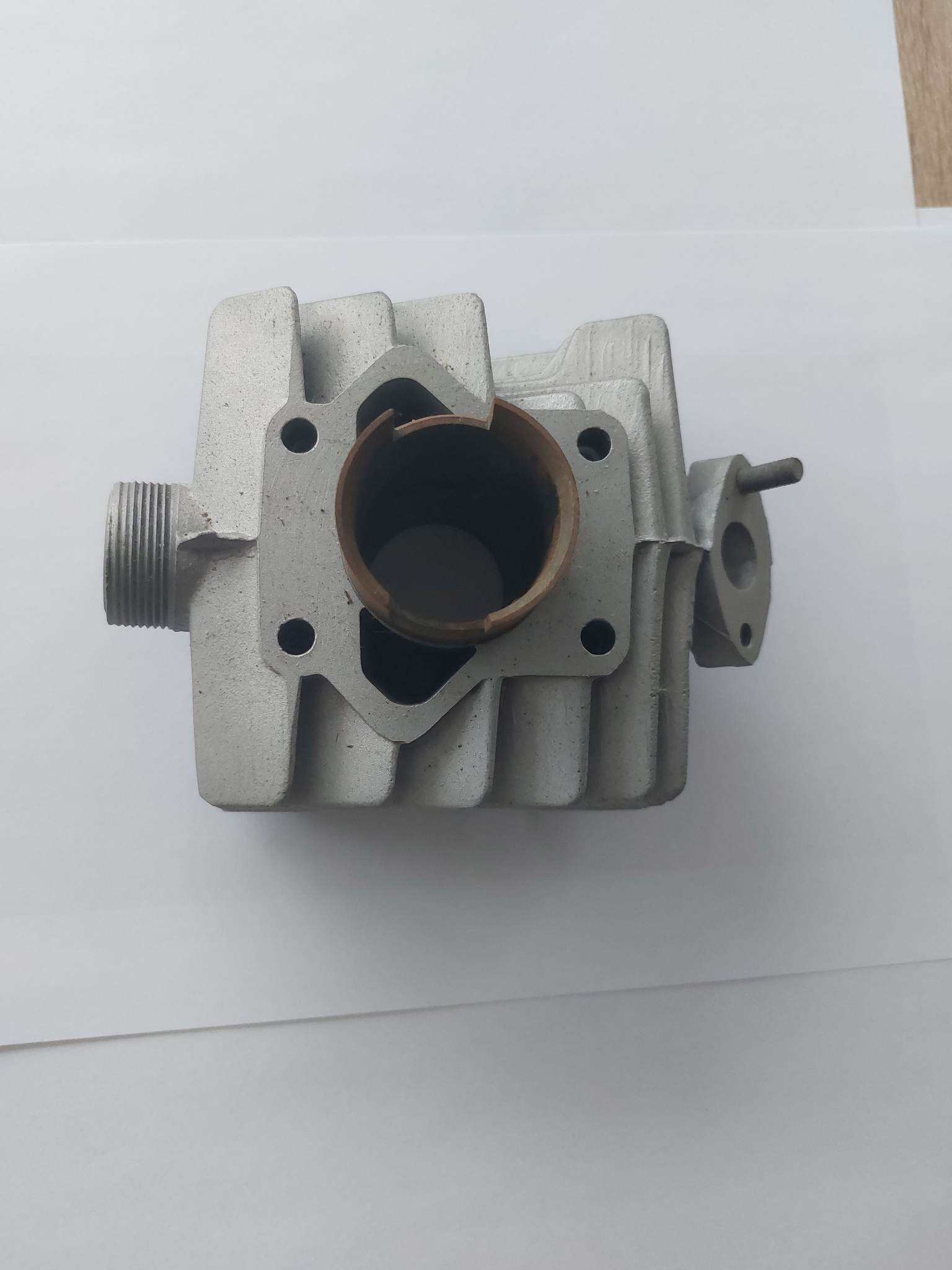 Szary, odlewany blok silnika z widocznymi otworami montażowymi i gwintowanym wlotem, leżący na białym tle.