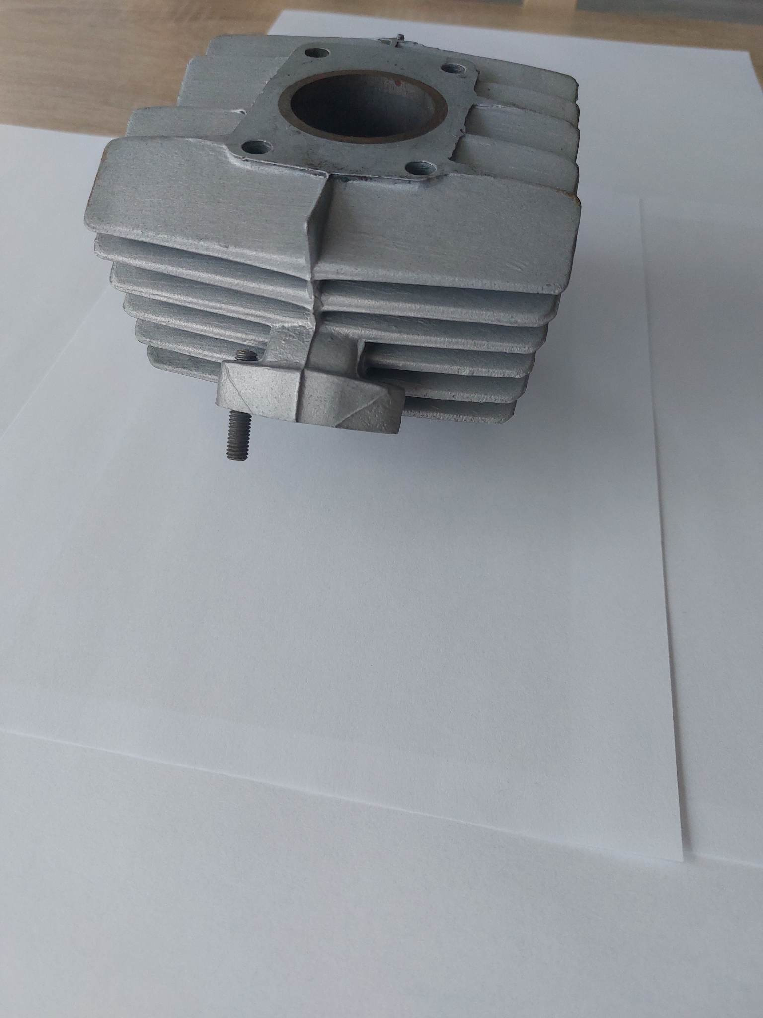 Szary cylinder silnika po piaskowaniu, widoczne ślady obróbki i gwintowany otwór, leżący na białym papierze.