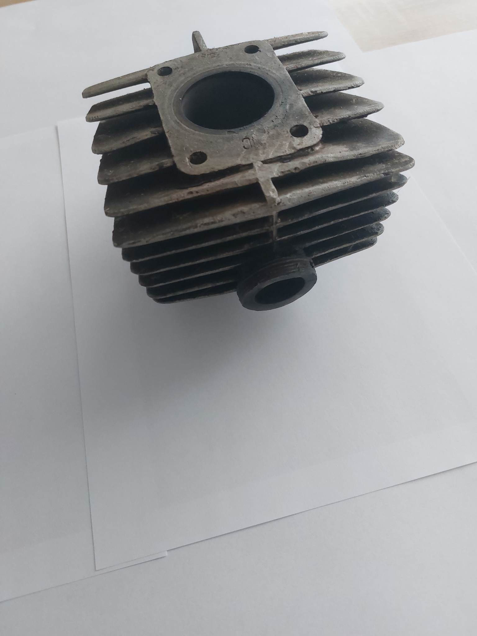 Cylindryczna, żeliwna głowica silnika spalinowego, widoczne żebra chłodzące, otwory montażowe i ślady korozji, umieszczona na białym tle.