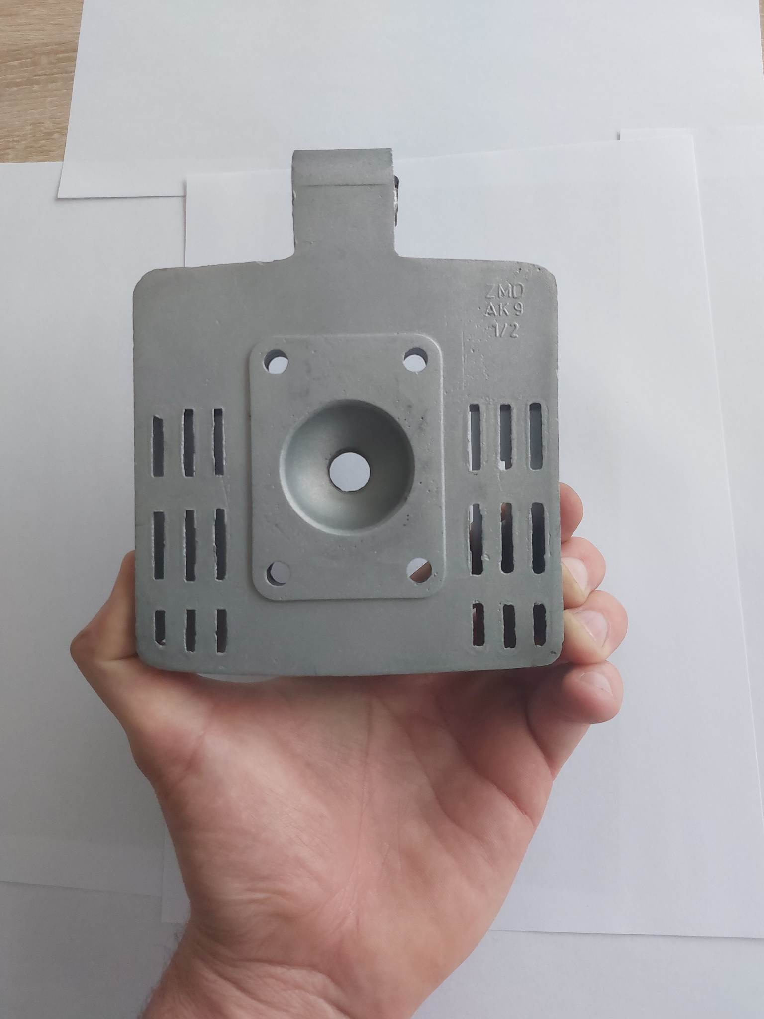 Szary, metalowy element, prawdopodobnie głowica silnika, trzymany w dłoni na białym tle, widoczne otwory i napisy ZMD AK9 1/2.