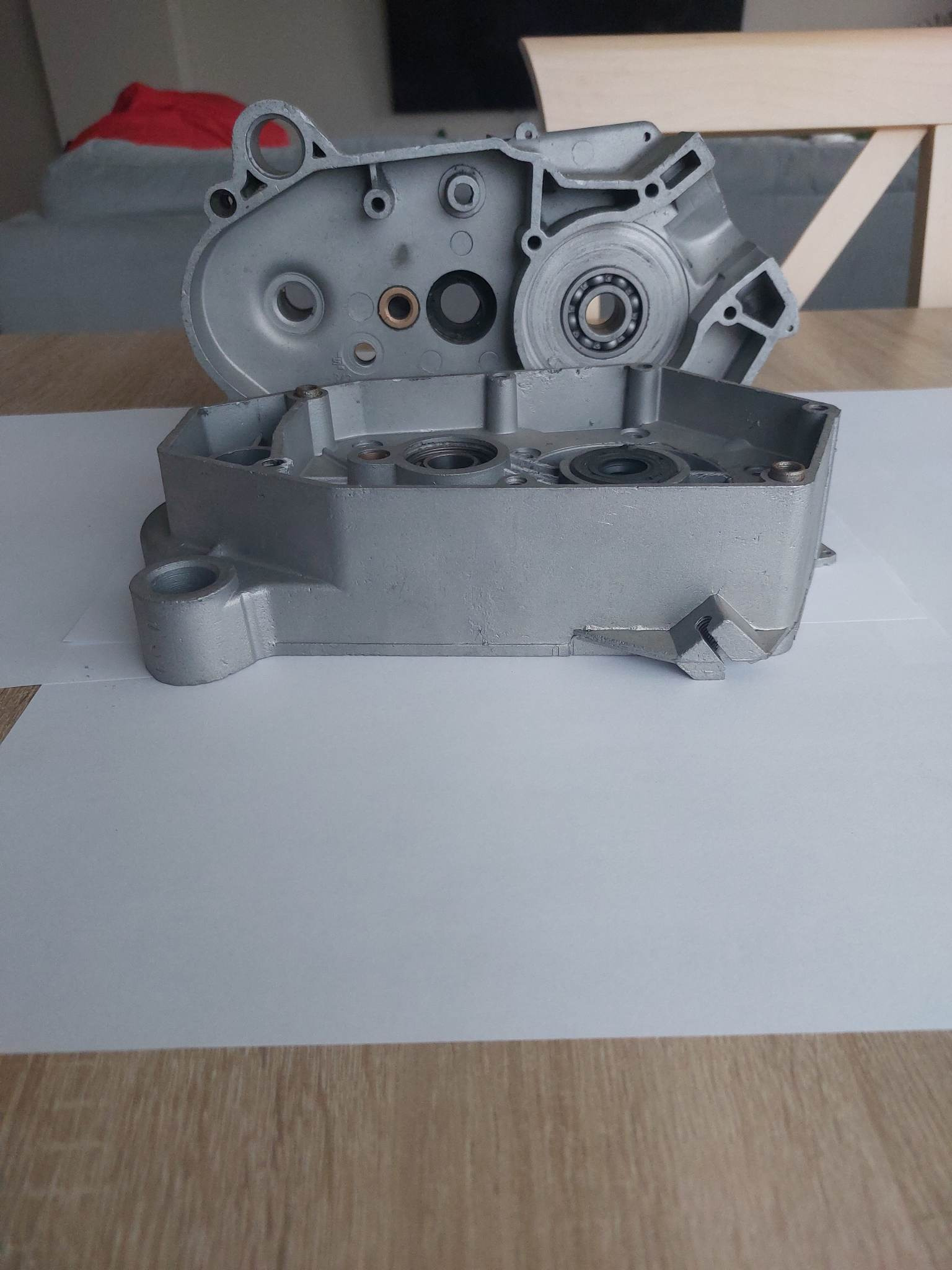 Dwie połówki aluminiowej obudowy silnika motocyklowego, pokryte srebrną farbą, leżące na białym papierze na drewnianym stole.