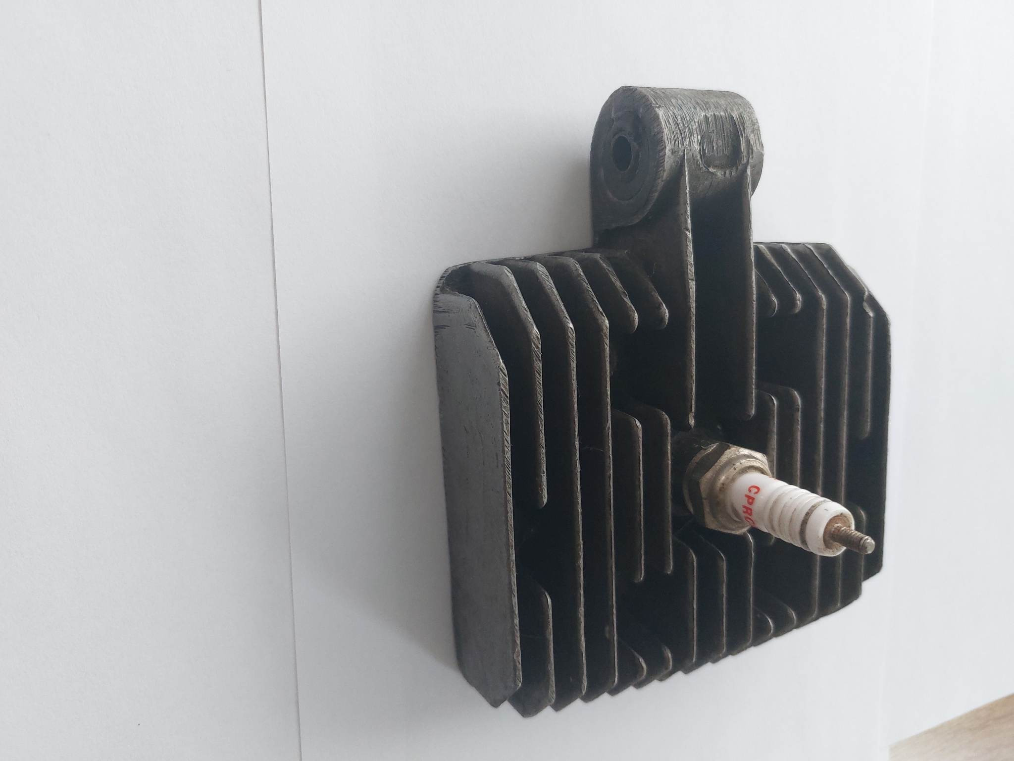 Szary, metalowy radiator z żebrami chłodzącymi i wystającą świecą zapłonową, umieszczony na białym tle.