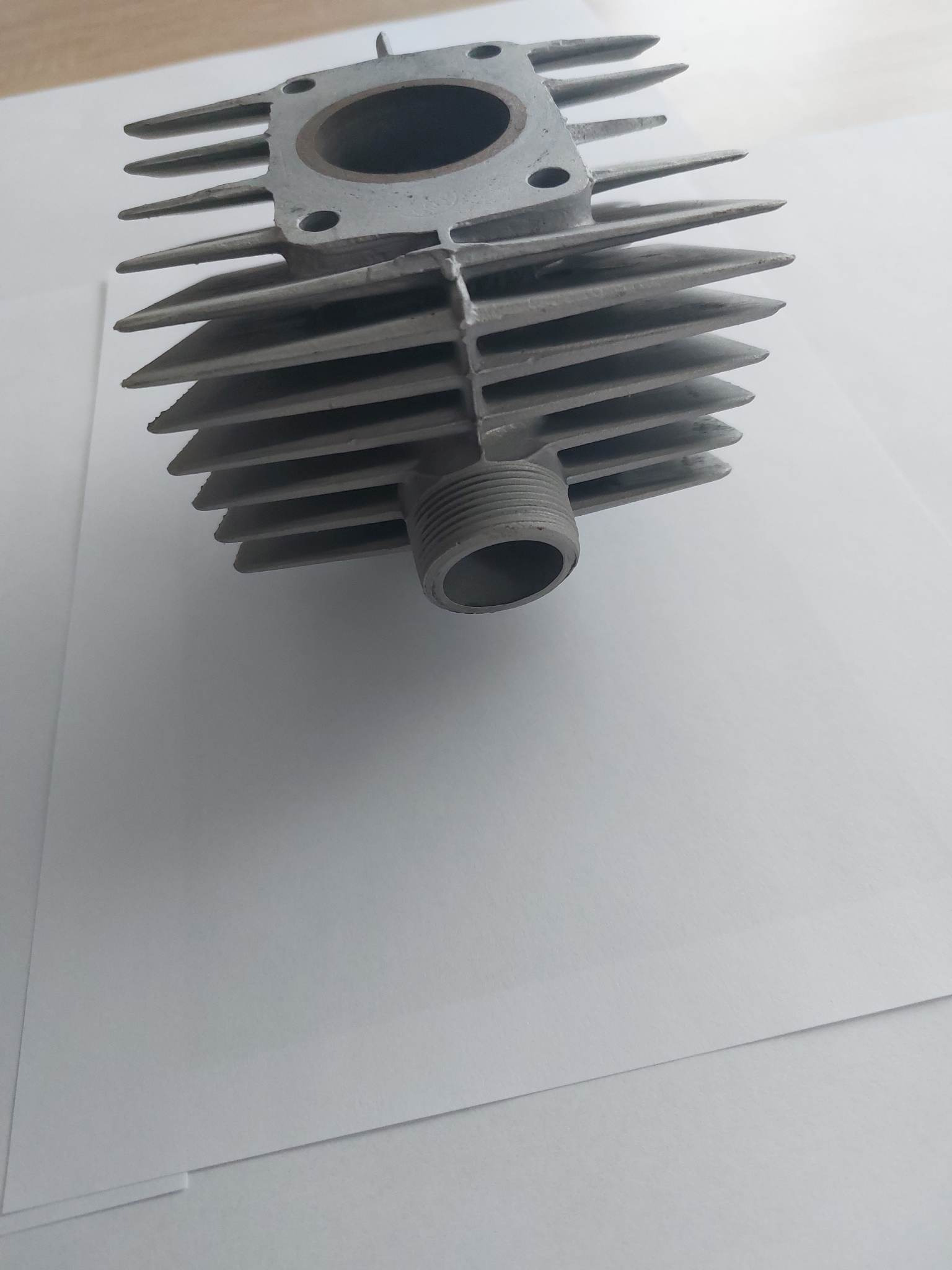 Szary, surowy blok cylindra silnika z aluminiowymi żebrami chłodzącymi, widoczne gwintowane wyjście, umieszczony na białym tle.