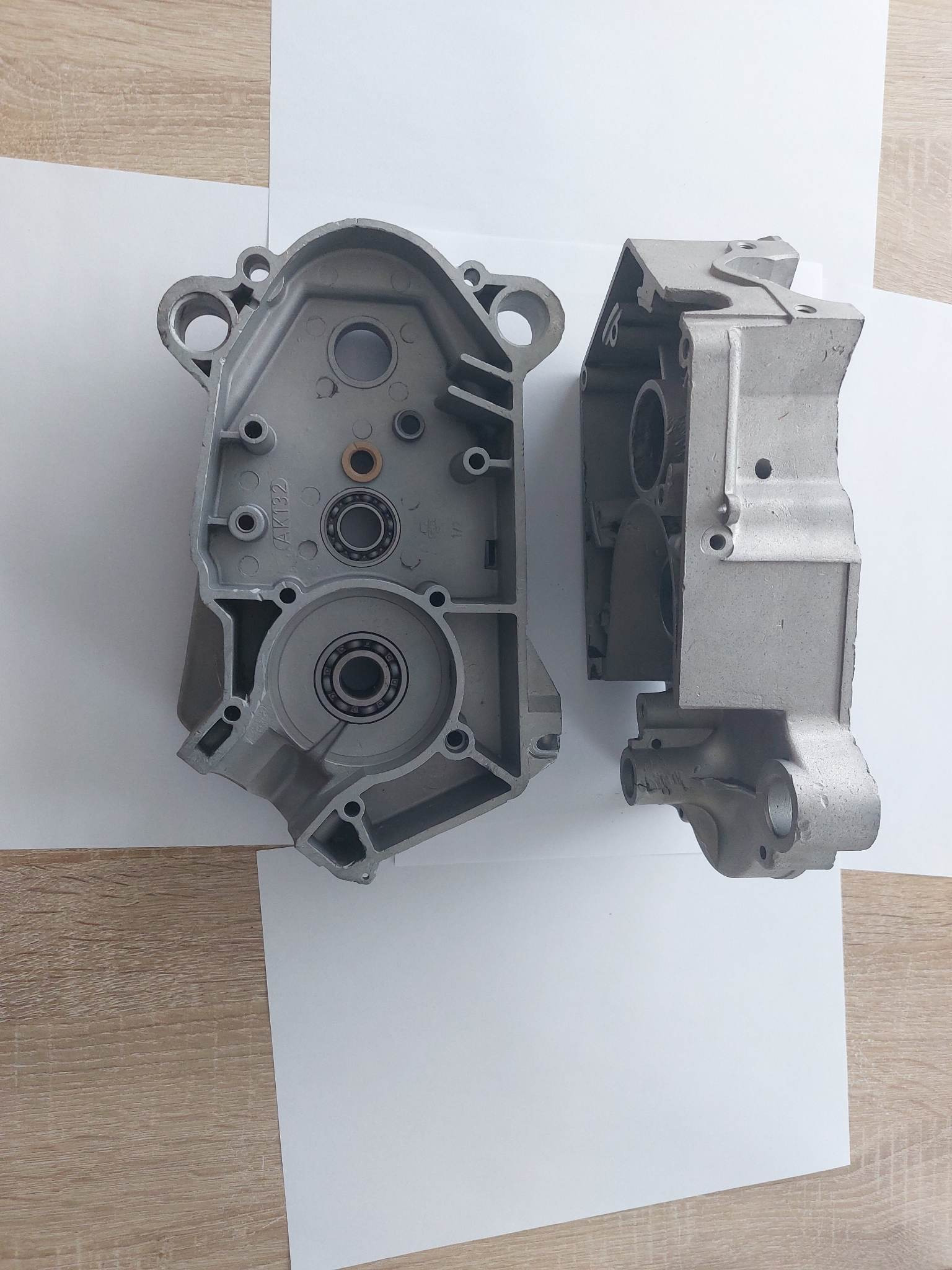 Dwie połówki aluminiowej obudowy silnika, widoczne łożyska, leżą na białym papierze na drewnianym blacie.