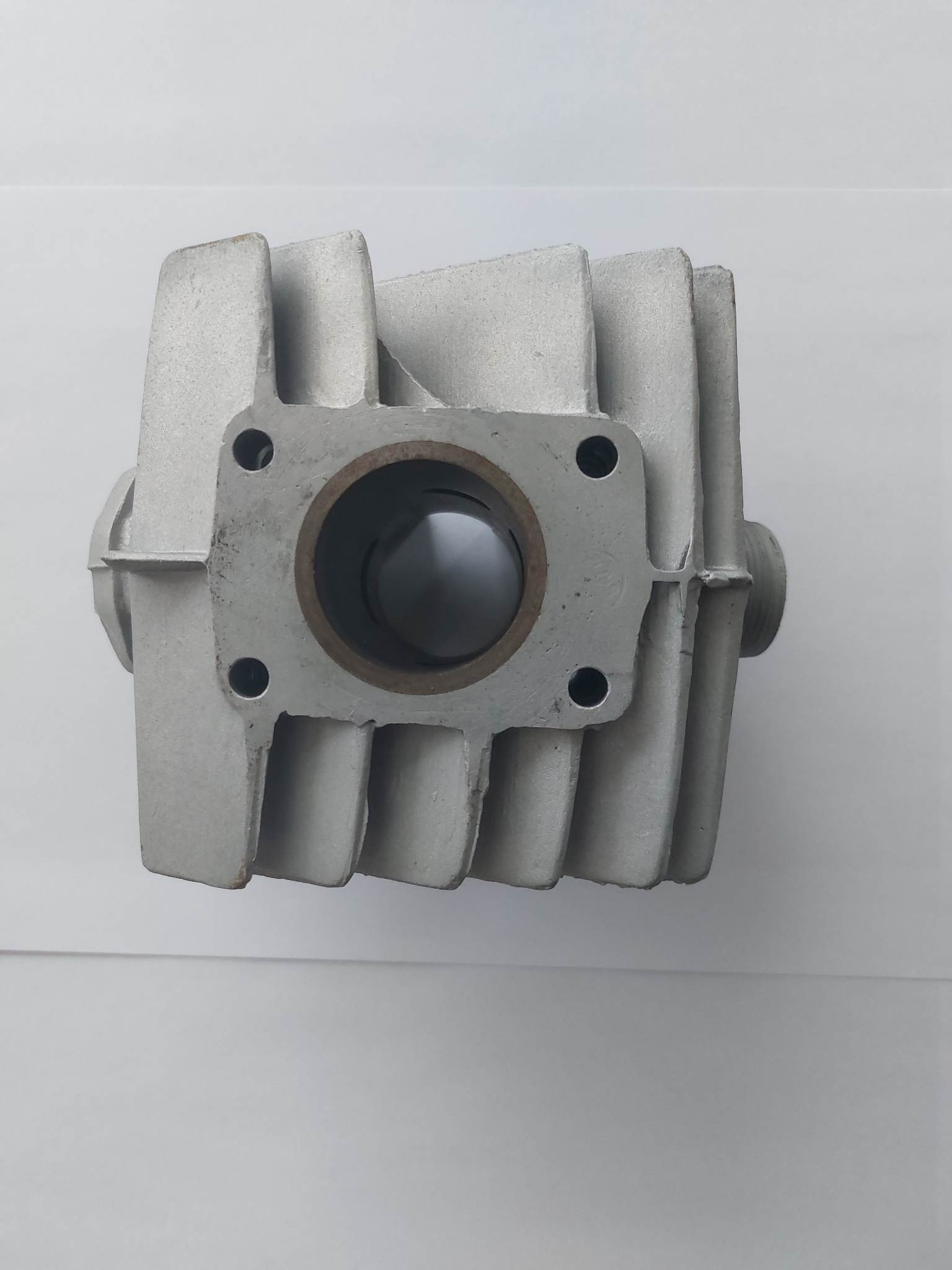 Surowy, odlewany aluminiowy cylinder silnika spalinowego z widocznymi żebrami chłodzącymi i otworami montażowymi, leżący na białym tle.