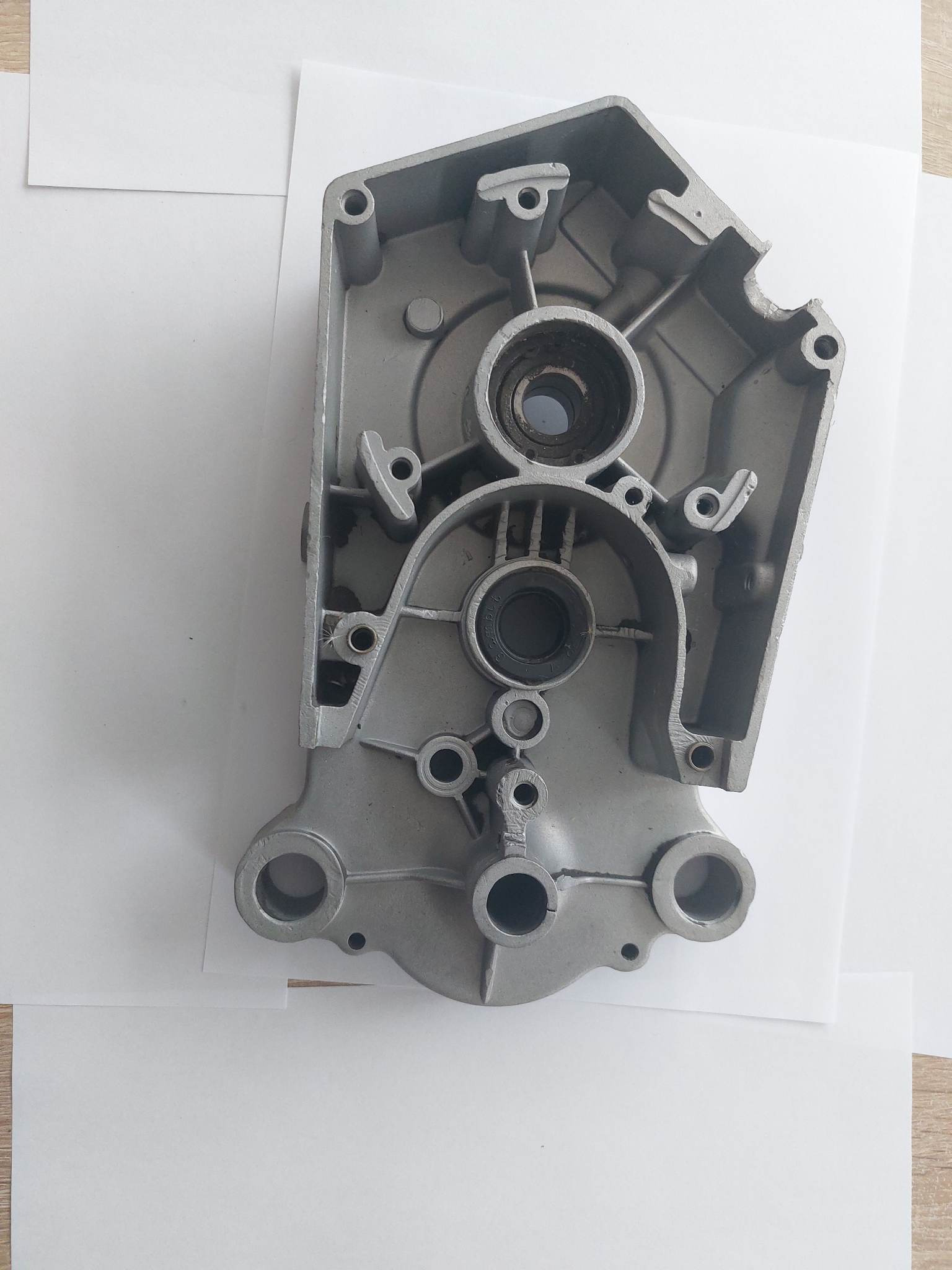 Szara, aluminiowa obudowa silnika z widocznymi otworami i mocowaniami, leżąca na białym papierze na drewnianym stole.