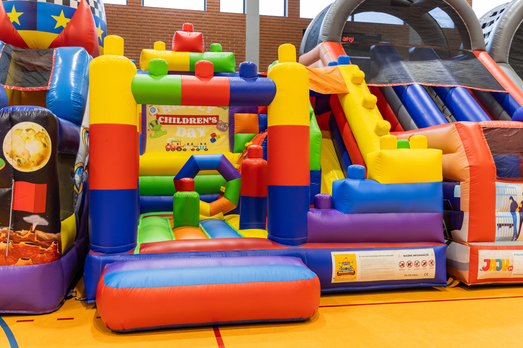 Kolorowy, rozbudowany dmuchany plac zabaw z motywem kosmicznym i napisem 'Children's Day', ustawiony w hali sportowej.