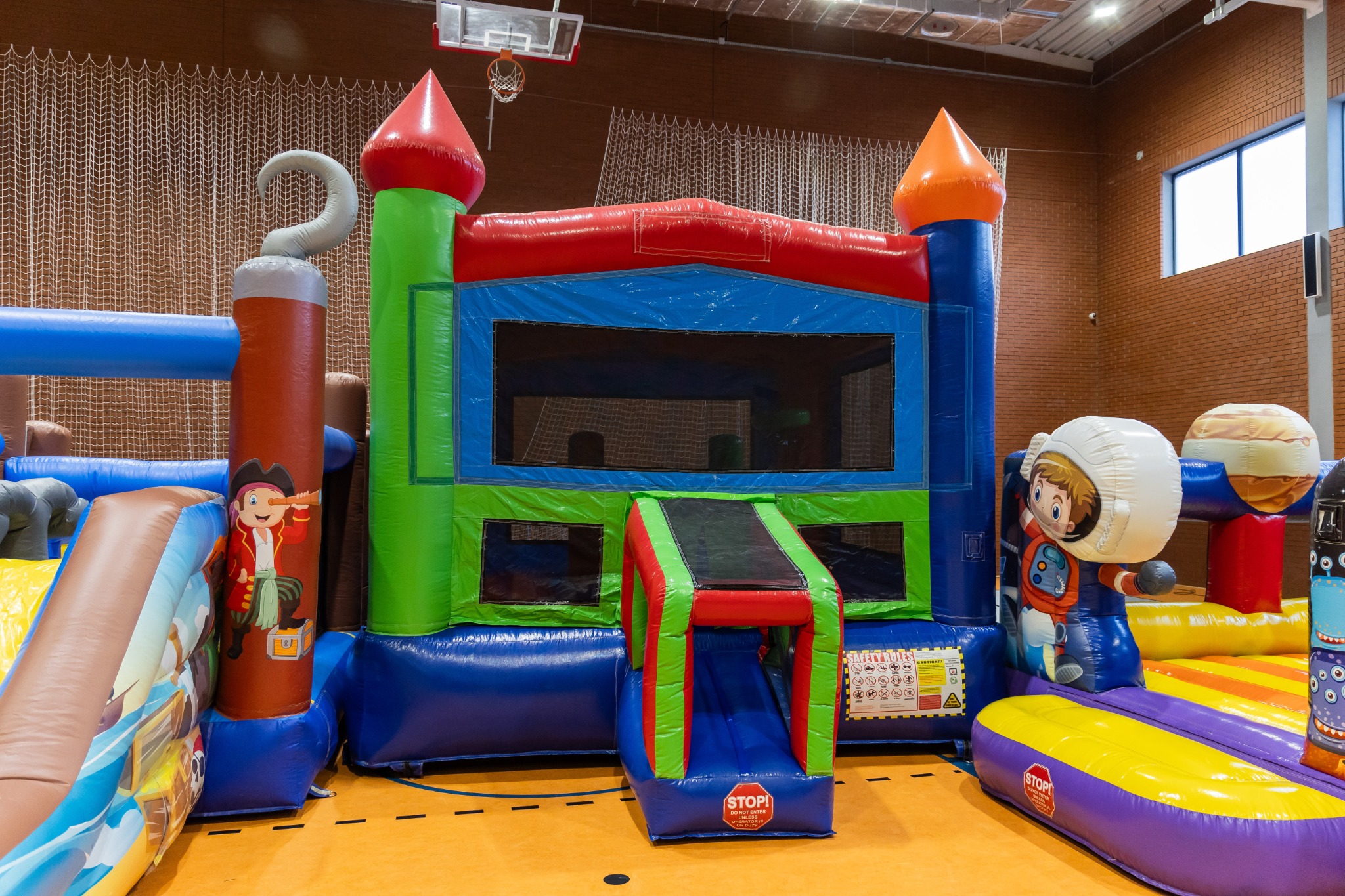 Kolorowy dmuchany zamek z motywami piratów i kosmosu, ustawiony w hali sportowej. Widoczny kosz do koszykówki w tle.