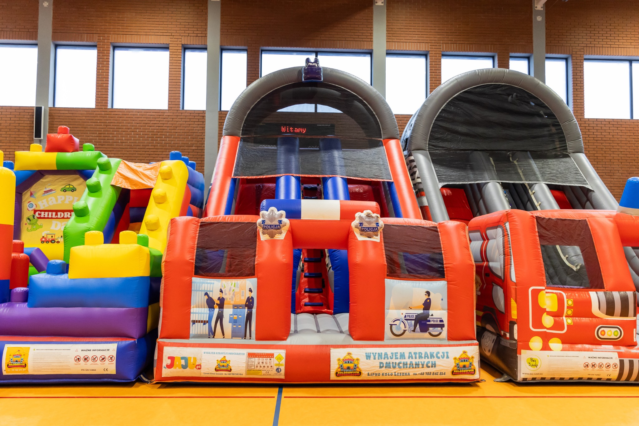 Kolorowe dmuchańce w hali sportowej, z motywami policyjnymi i dziecięcymi, ustawione obok siebie, z widocznymi oknami i ceglaną ścianą w tle.