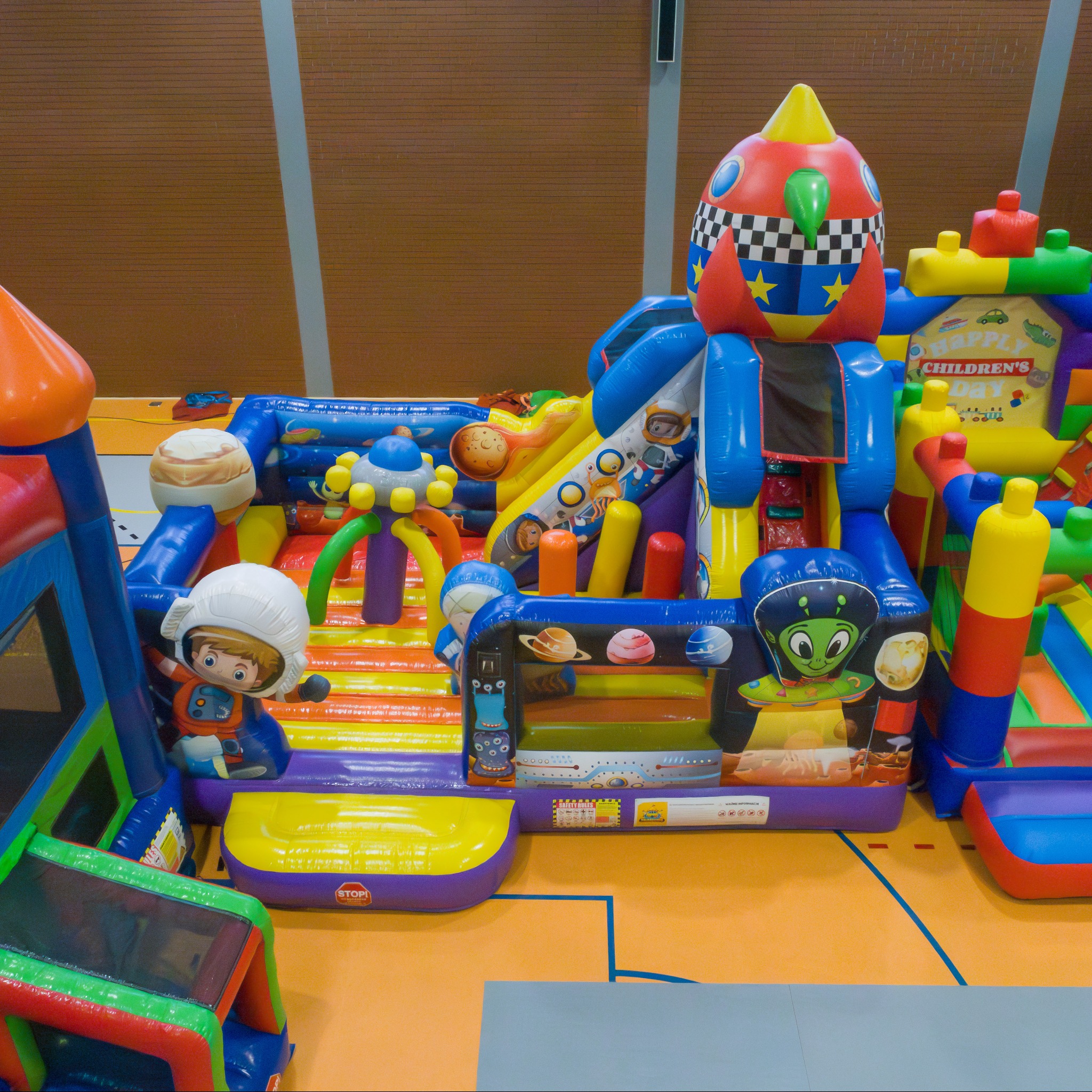 Kolorowy, rozbudowany dmuchany plac zabaw w motywie kosmicznym z rakietą, planetami i postacią astronauty, ustawiony w hali sportowej.