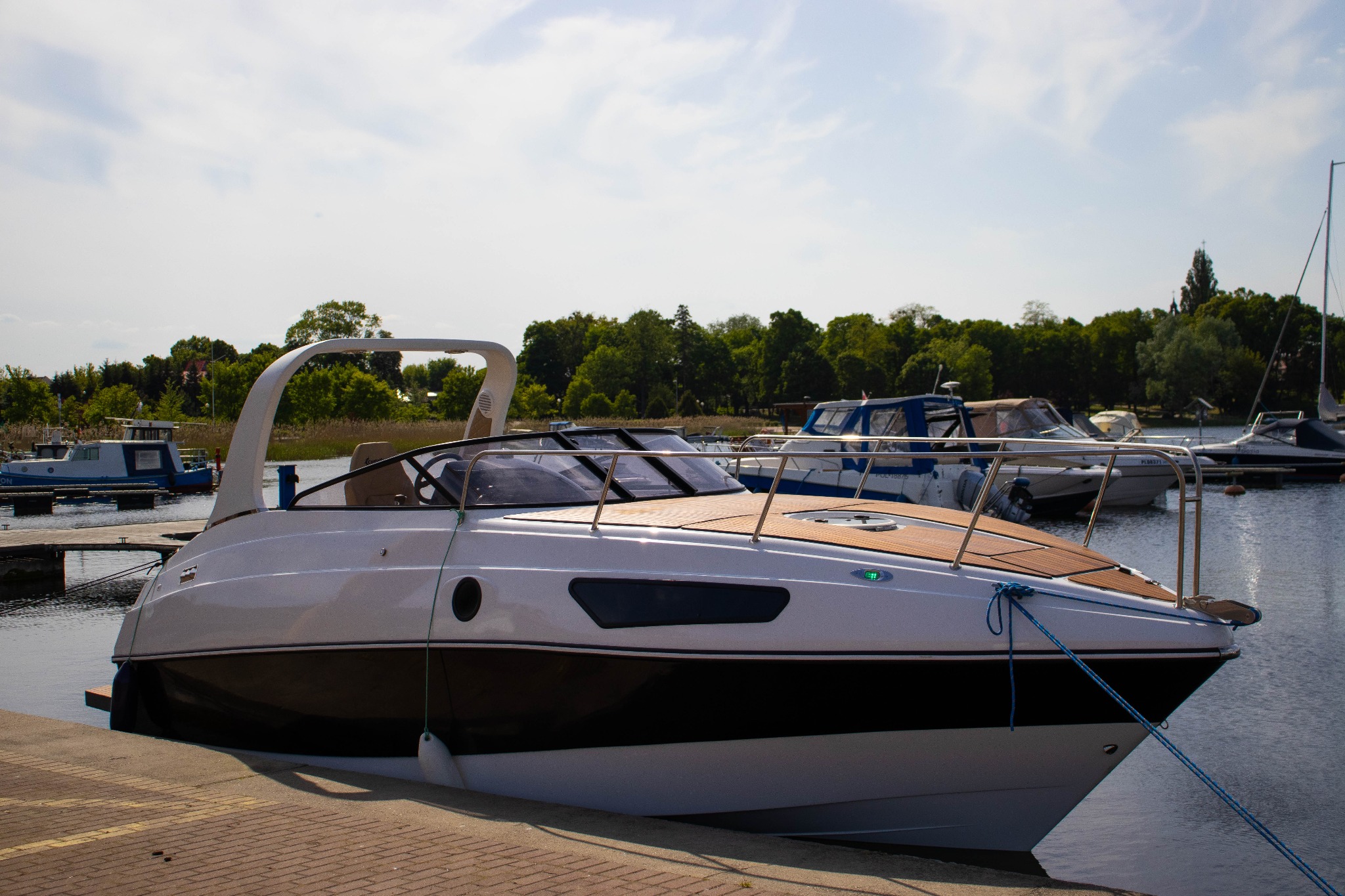 Biała motorówka cumująca przy ceglanym nabrzeżu, z widocznymi innymi łodziami i drzewami w tle, niebieska lina cumownicza zabezpiecza łódź.