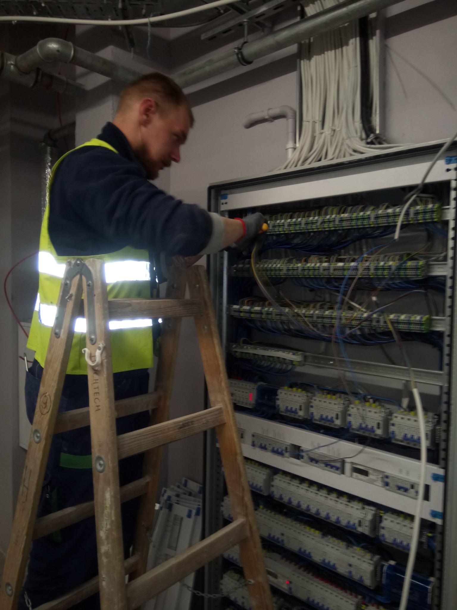 Elektryk w kamizelce odblaskowej na drewnianej drabinie, pracujący przy skrzynce bezpiecznikowej z licznymi przewodami i elementami elektrycznymi w pomieszczeniu technicznym.