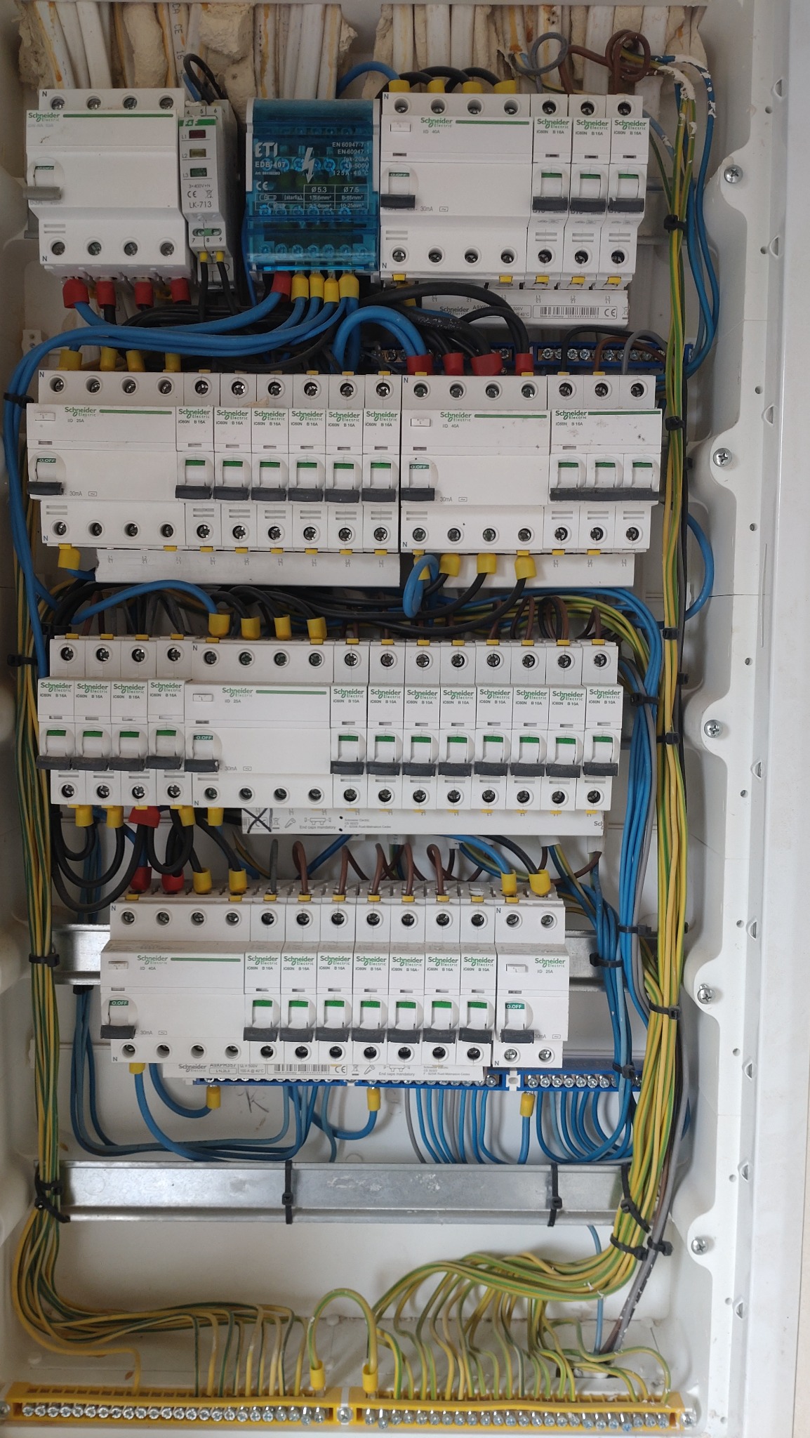 Rozdzielnica elektryczna z zainstalowanymi bezpiecznikami Schneider Electric oraz przewodami w kolorach niebieskim, żółto-zielonym i czarnym, uporządkowane wewnątrz białej obudowy.