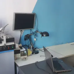 Stanowisko pracy z lutownicą BGA, monitorem, lampką i otwartym laptopem; biało-niebieskie tło.