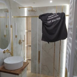 JSC Rafał Simkowski - Nowoczesna łazienka po remoncie z kabiną prysznicową, umywalką nablatową i złotymi akcentami, częściowo zasłonięta czarną koszulką z nadrukiem 'Remonty & Wykończenia' i numerem telefonu.