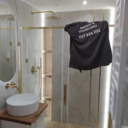 Nowoczesna łazienka po remoncie z kabiną prysznicową, umywalką nablatową i złotymi akcentami, częściowo zasłonięta czarną koszulką z nadrukiem 'Remonty & Wykończenia' i numerem telefonu.