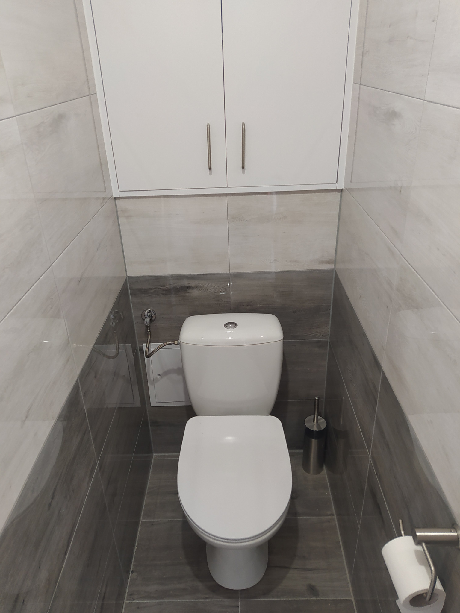 Nowoczesna toaleta w szaro-białej łazience z wiszącą szafką nad nią, widoczny pojemnik na szczotkę do WC i uchwyt na papier toaletowy.