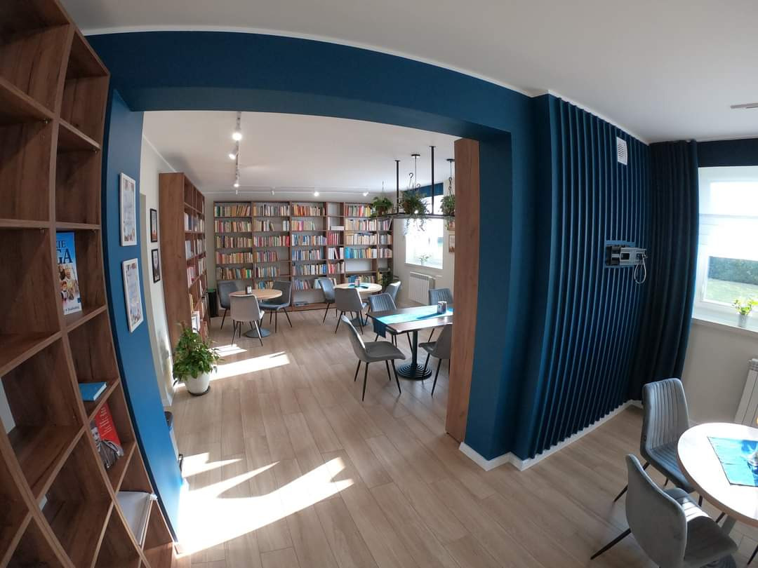 Przestronne wnętrze biblioteki z regałami pełnymi książek, stolikami i krzesłami dla czytelników, drewnianą podłogą i akcentami w kolorze głębokiego błękitu.