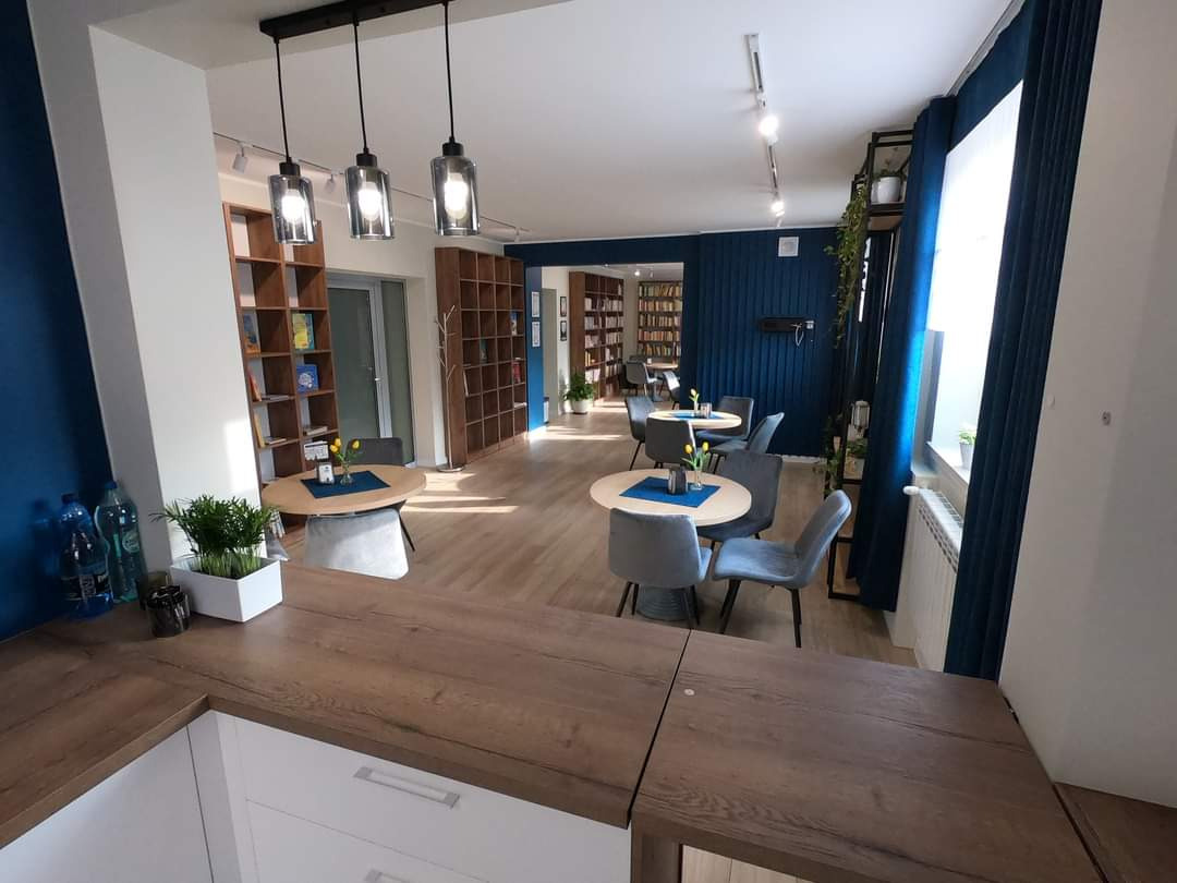 Jasne wnętrze kawiarni lub biblioteki z drewnianymi regałami, okrągłymi stolikami z szarymi krzesłami i błękitnymi akcentami kolorystycznymi na ścianach i zasłonach.