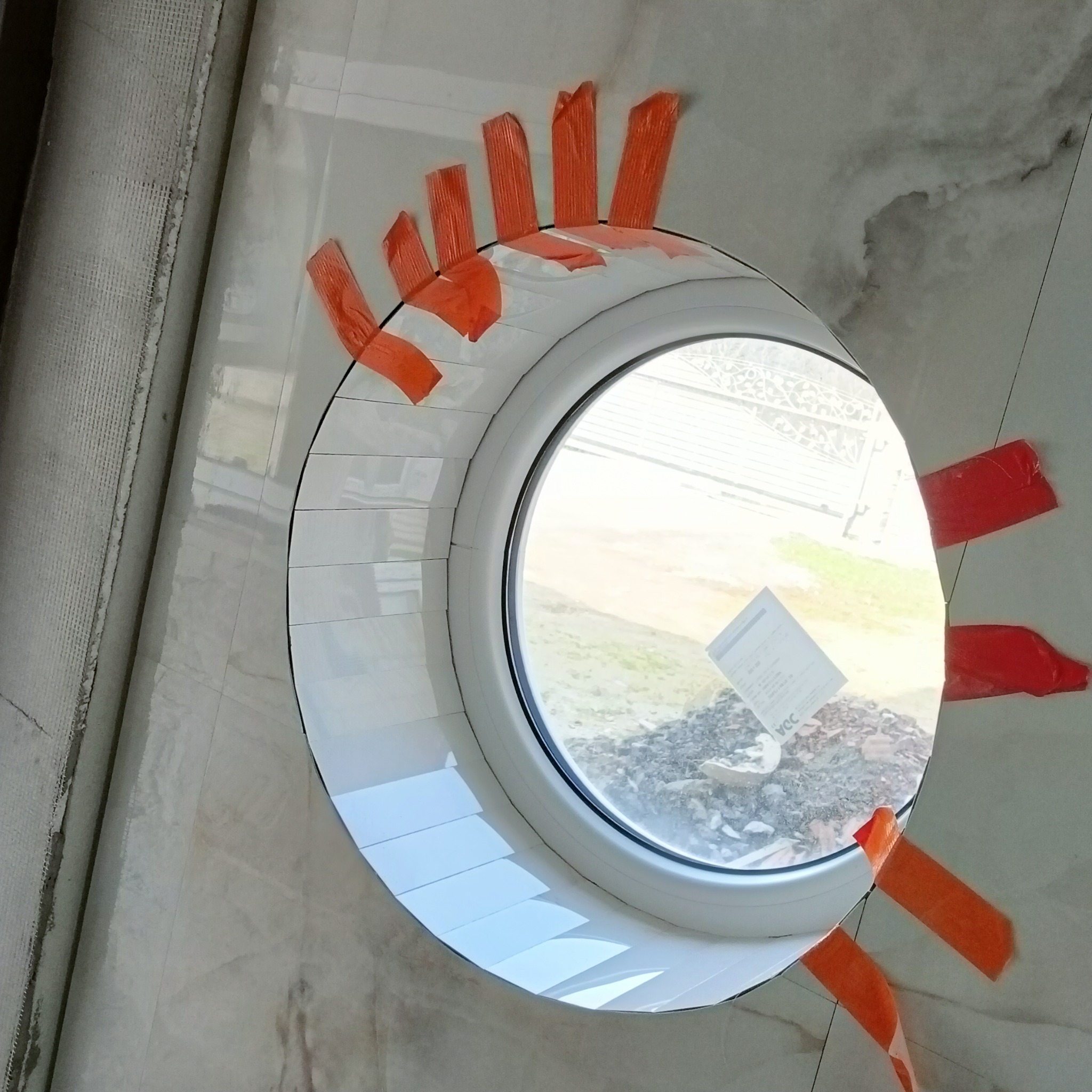 Okrągłe okno obłożone białymi płytkami, zabezpieczone pomarańczową taśmą malarską, widoczne niedociągnięcia w ułożeniu płytek i zabrudzenia na ścianie.