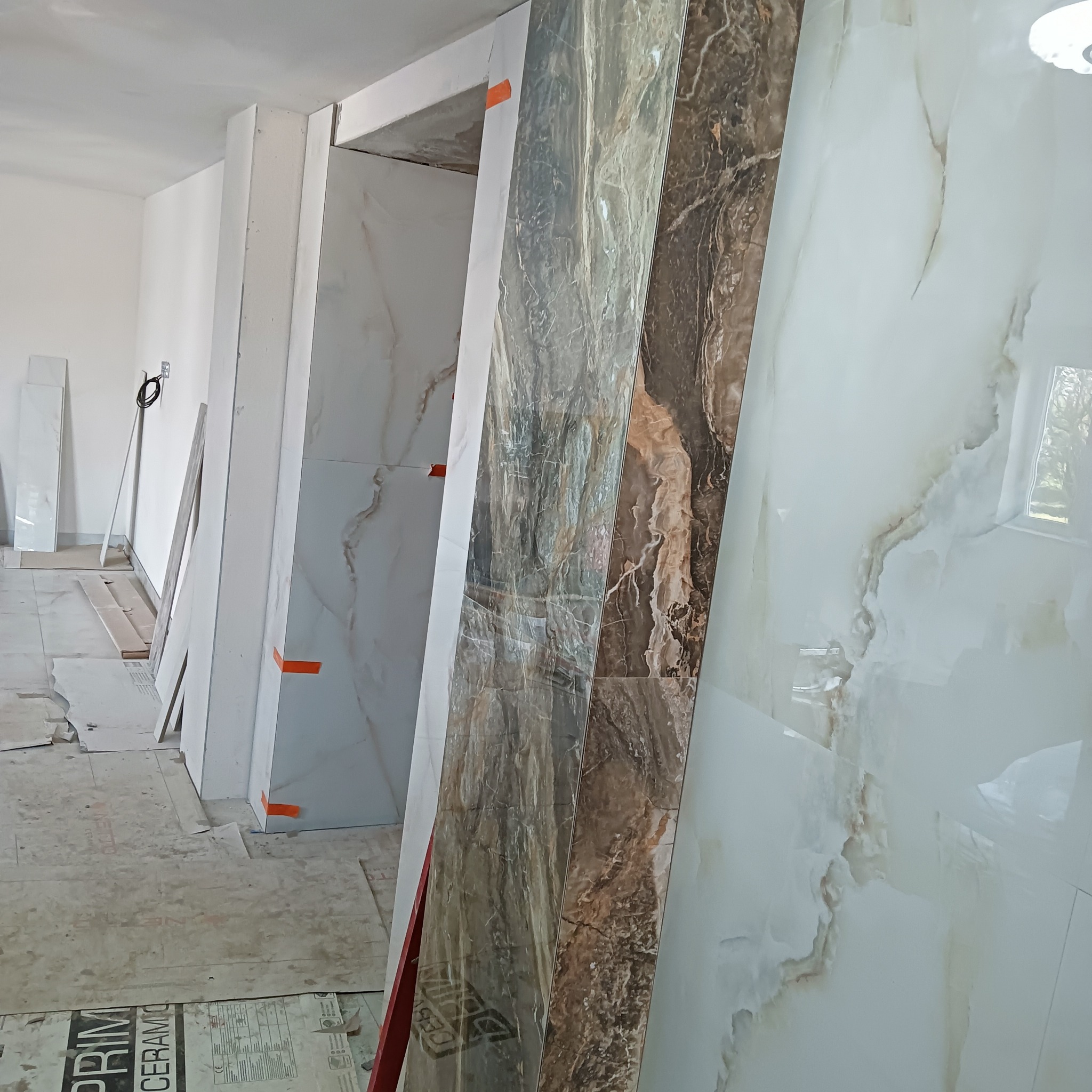 Wnętrze w trakcie prac: ułożone przy ścianach płytki imitujące marmur w odcieniach bieli i brązu, zabezpieczone pomarańczową taśmą, surowy stan wykończenia, widoczne kable i podłoga zabezpieczona...