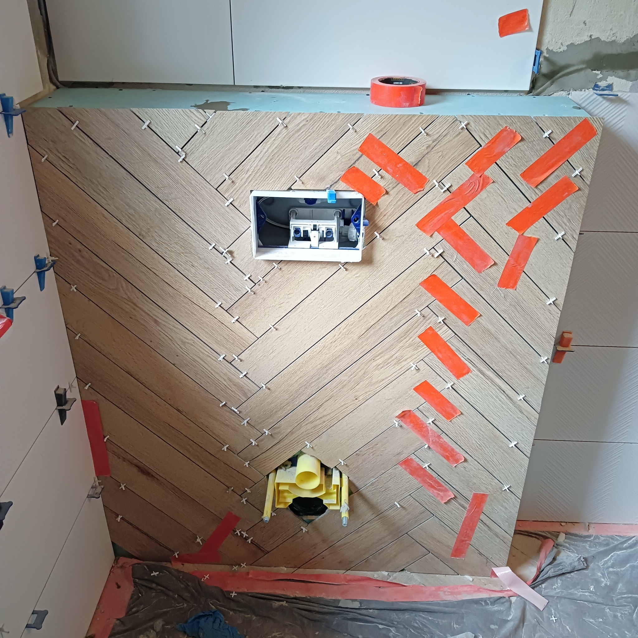 Ściana w trakcie układania płytek imitujących drewno w jodełkę, z widocznymi elementami instalacji sanitarnych, taśmą malarską i krzyżykami dystansowymi.