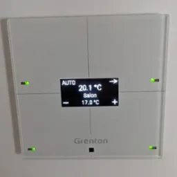 Biały panel sterowania Grenton Smart Home zamontowany na ścianie, wyświetlający temperaturę w salonie (20.1°C) oraz temperaturę docelową (17.0°C), z aktywnymi zielonymi diodami LED w rogach.